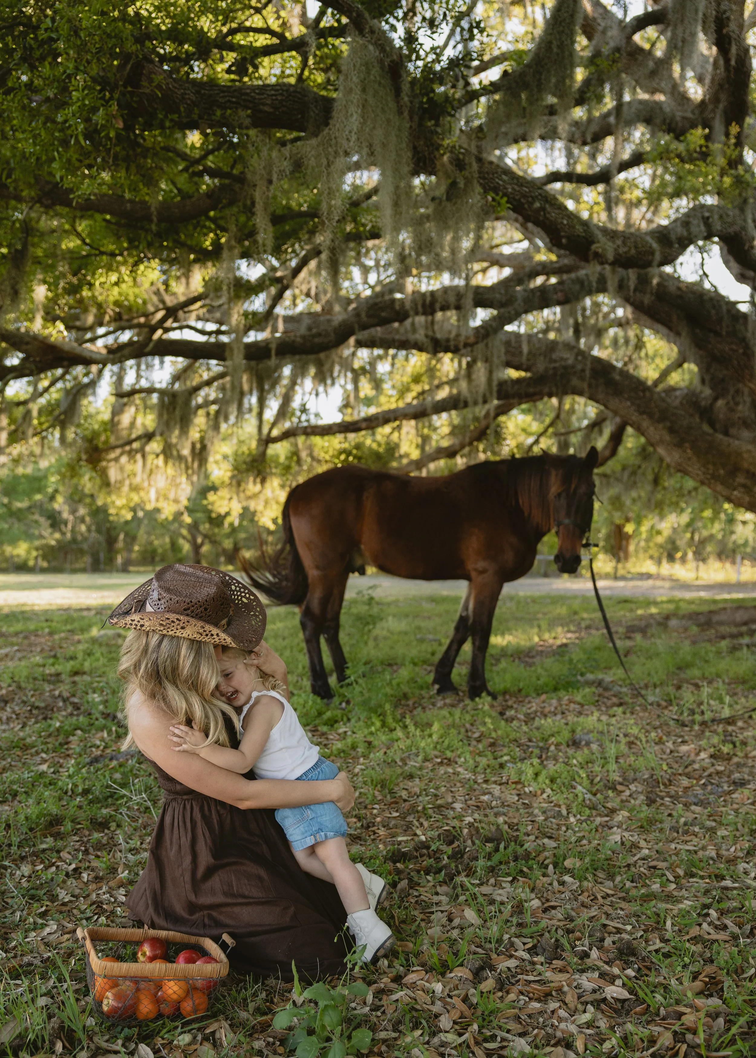 A woman holding two young children in an outdoor setting under a large tree with hanging moss. The woman is sitting on the ground, embracing the children. There is a basket of apples on the ground nearby. In the background, a horse is grazing, and th