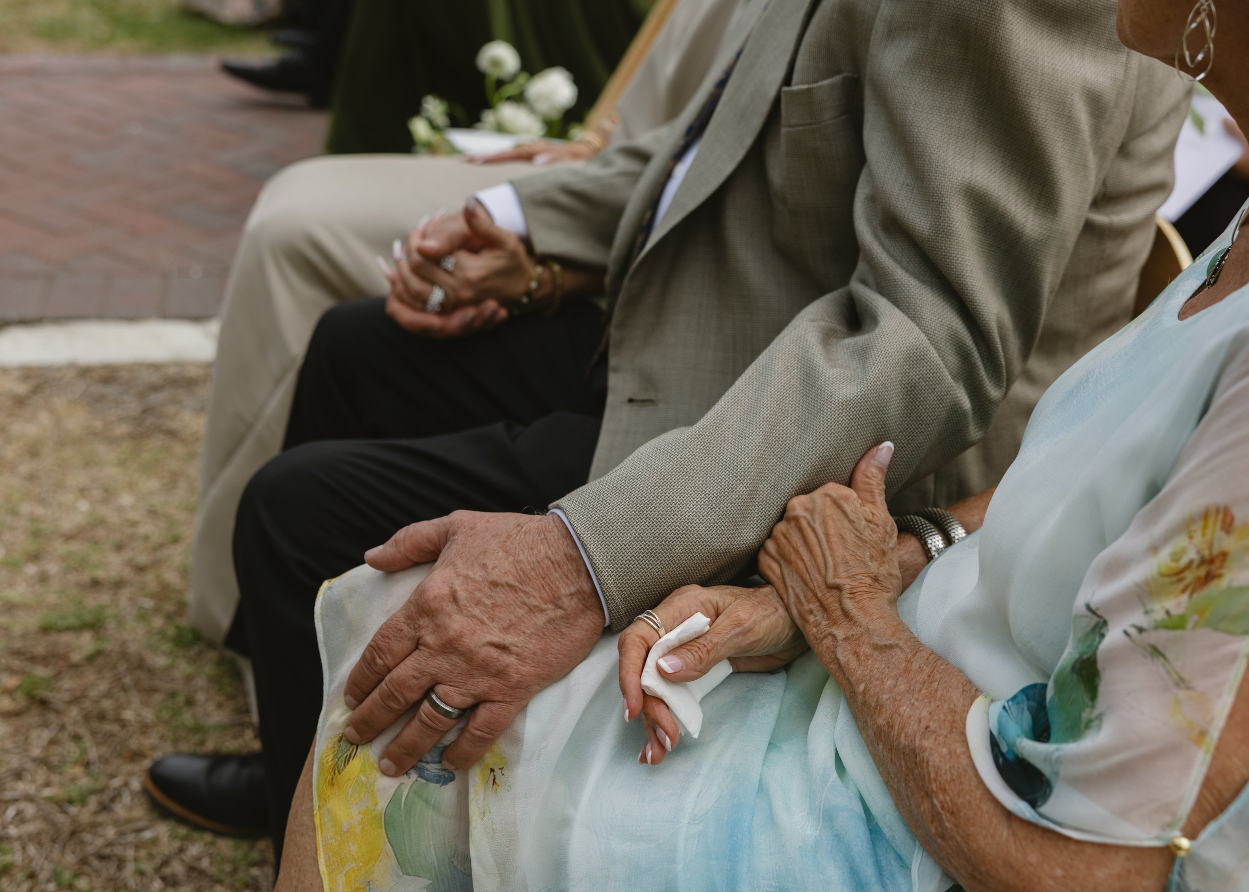 Close-up of elderly and middle-aged women holding hands during a gathering outdoors, with seating and flowers visible.