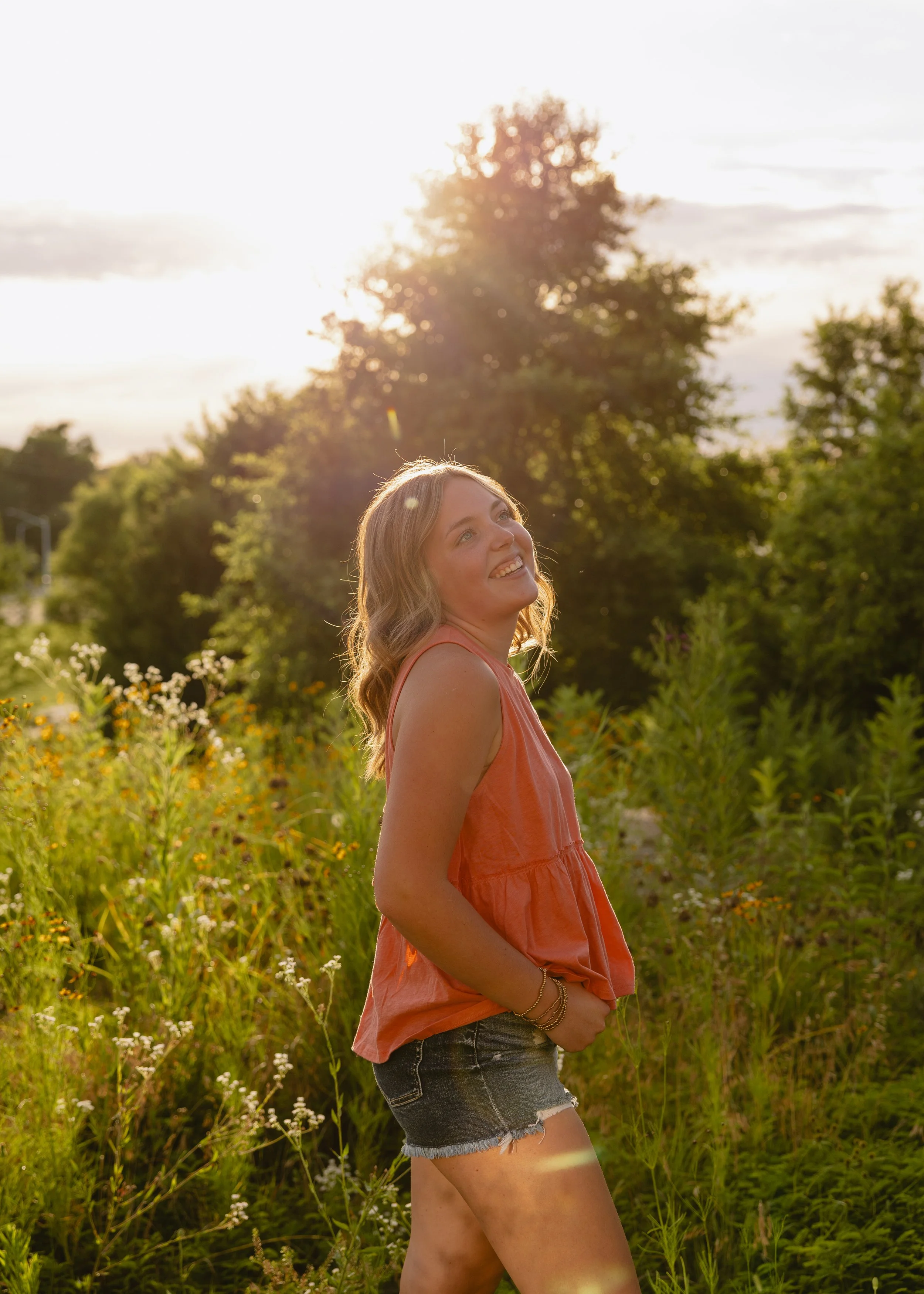 A young woman with wavy blonde hair and a big smile is standing in a field of tall green plants and flowers. She's wearing a sleeveless coral top, denim shorts, and has bracelets on her right wrist. The sun is setting behind a large tree, casting a w