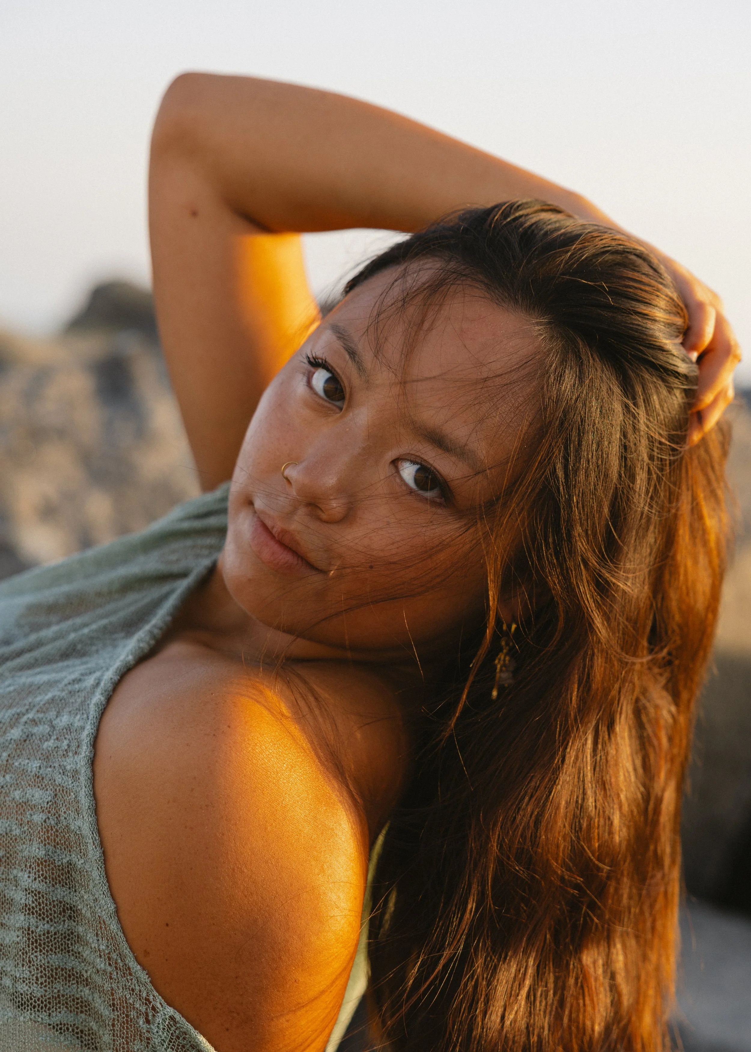 A young woman with long, wavy hair and a nose ring, outdoors during sunset, with her arm raised behind her head, looking at the camera.