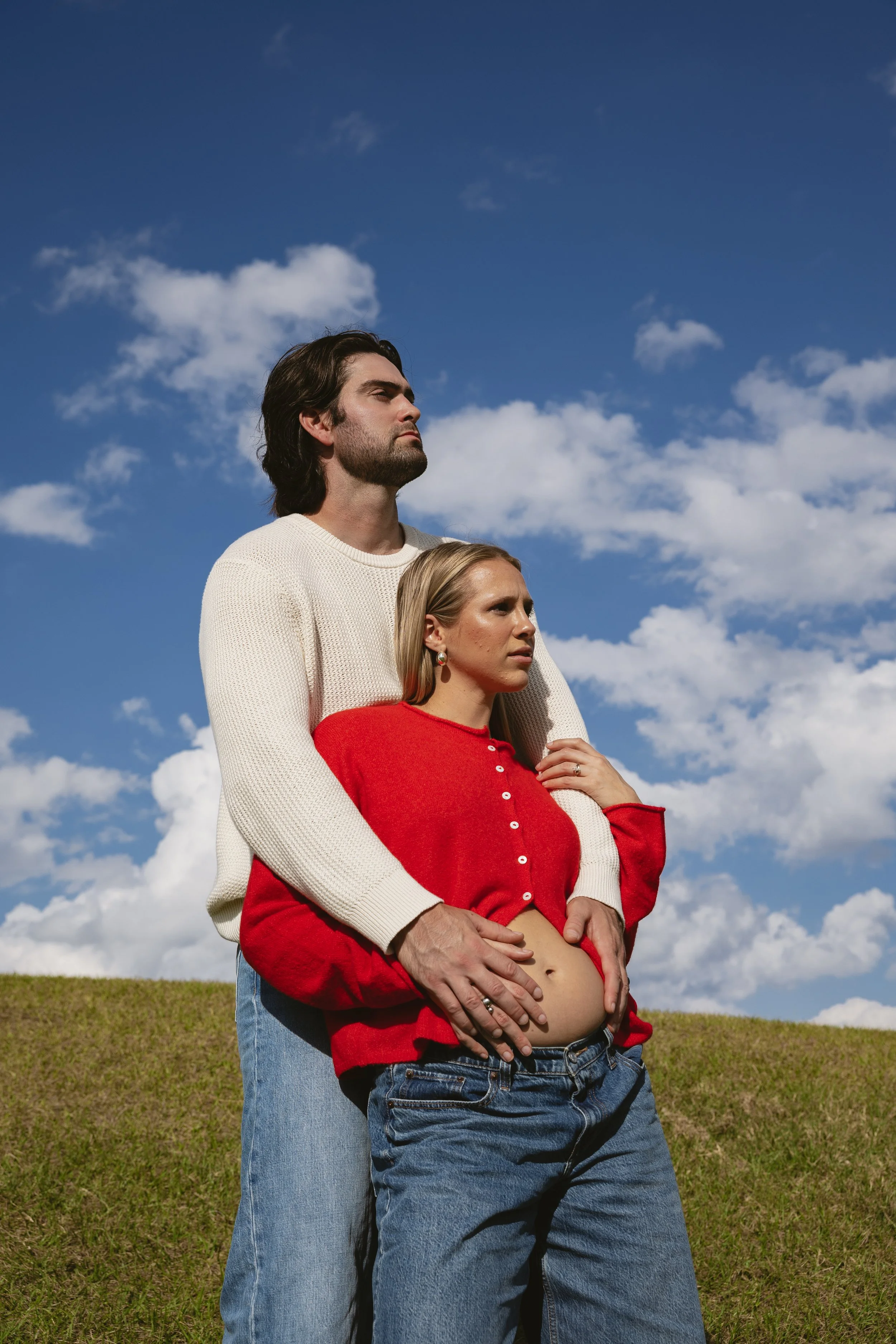 A man and a woman standing outdoors in a grassy field under a blue sky with scattered clouds. The woman is pregnant and is holding her belly, with the man standing behind her, both looking serious.