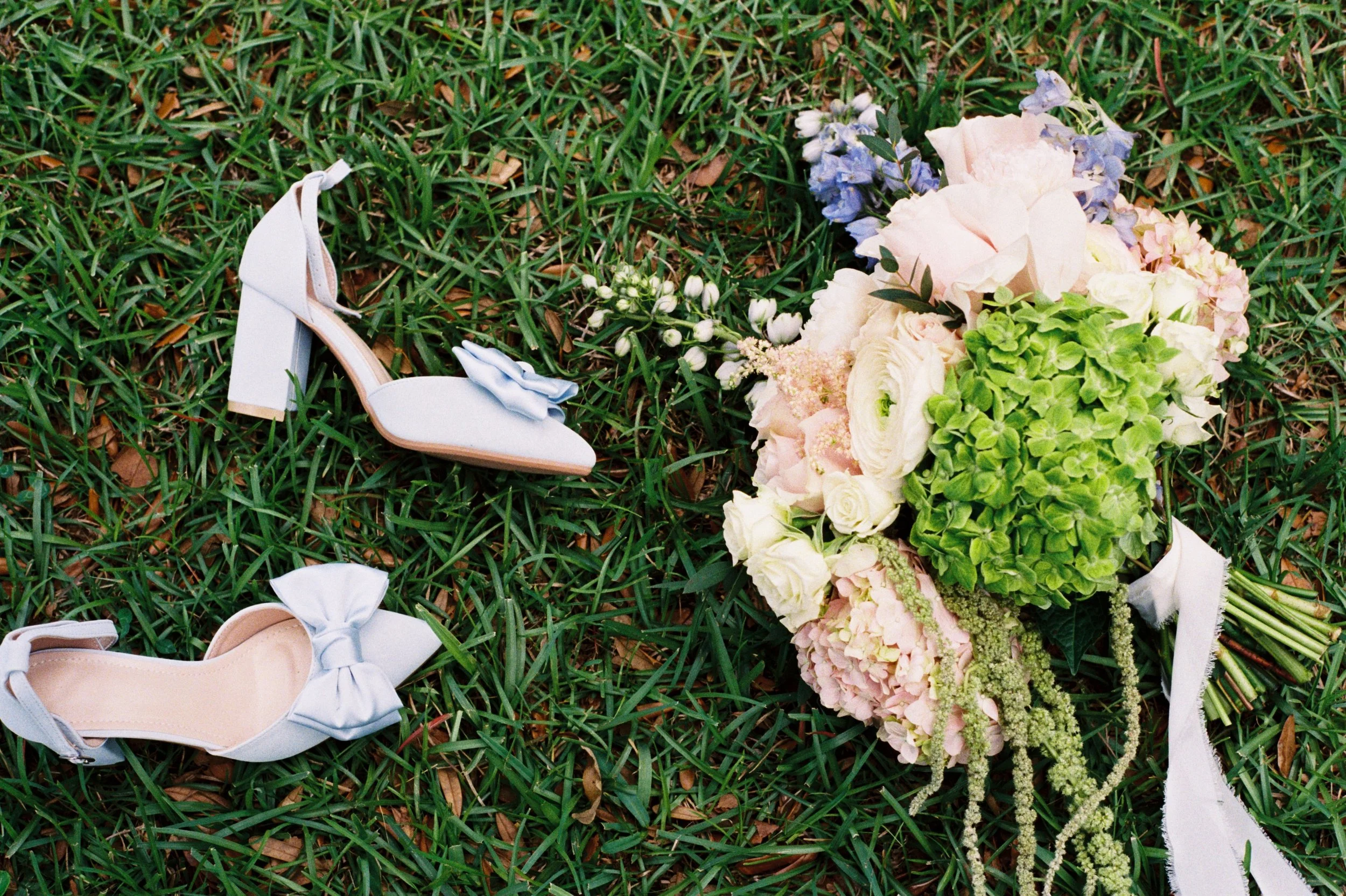 A white high heel shoe with a bow detail on the toe lying on green grass next to a bouquet of white, pink, and purple flowers.