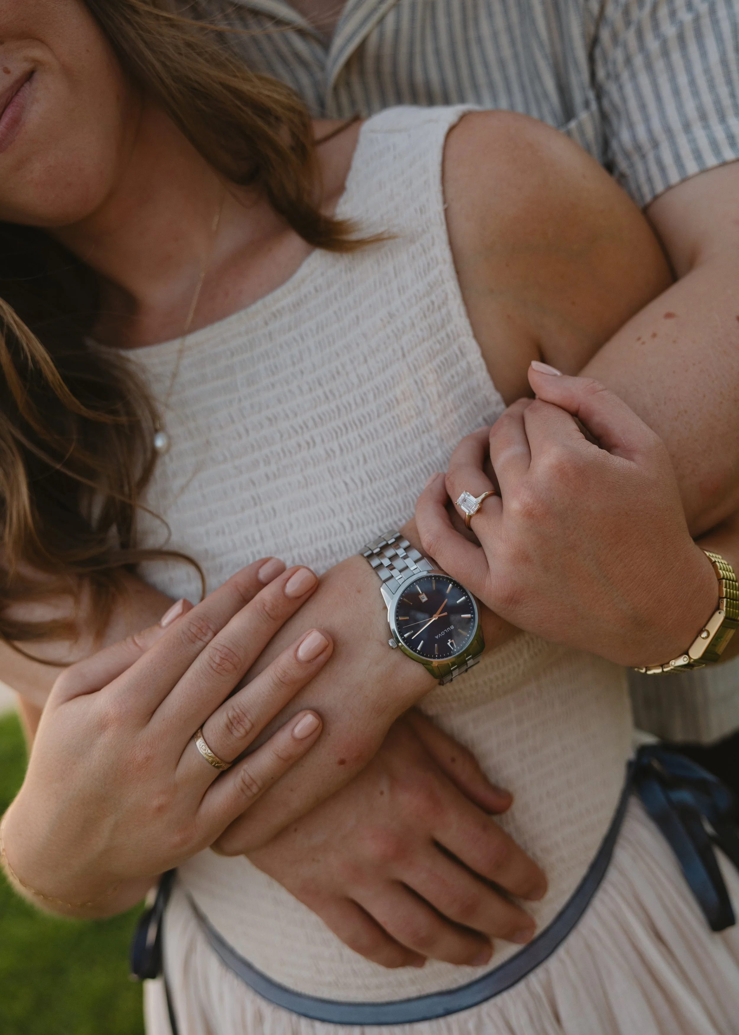 A couple embracing outdoors, with the woman's hand on the man's arm, showing a wedding ring, and the man's hand around the woman's waist, wearing a watch and bracelet. Both are dressed casually.