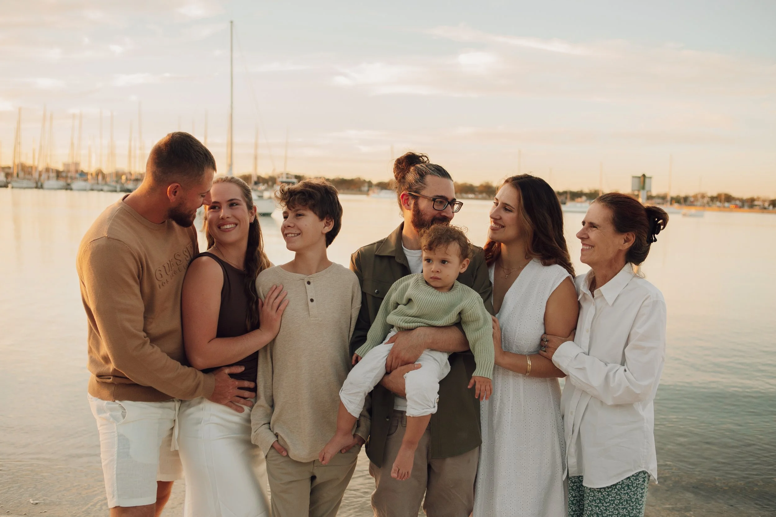 A multigenerational family of eight people standing by water at sunset, smiling and looking at each other, with sailboats and a marina in the background.