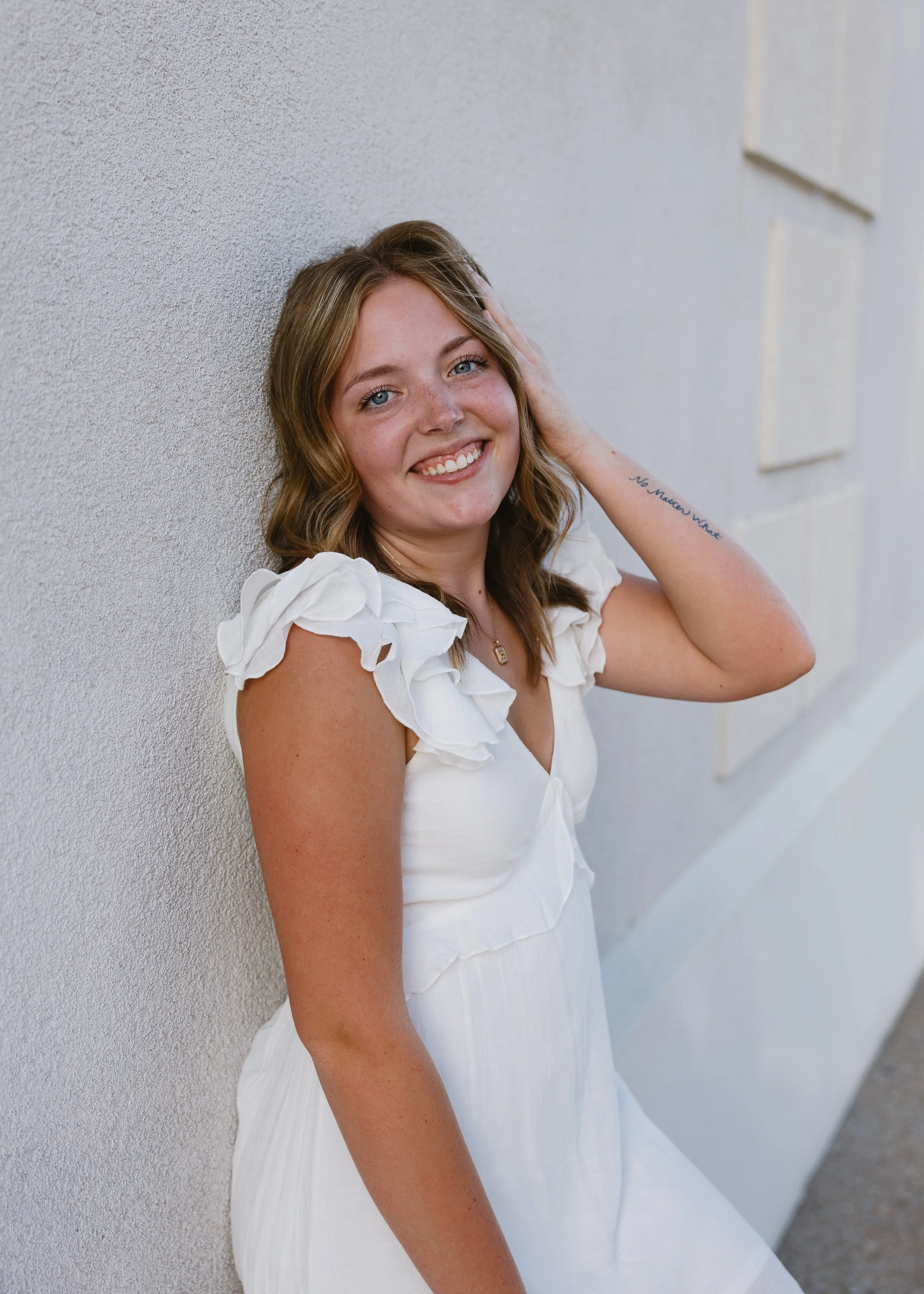 Smiling young woman with wavy light brown hair and blue eyes leaning against a textured white wall, wearing a white dress with ruffled sleeves, touching her hair with her right hand, and smiling at the camera.