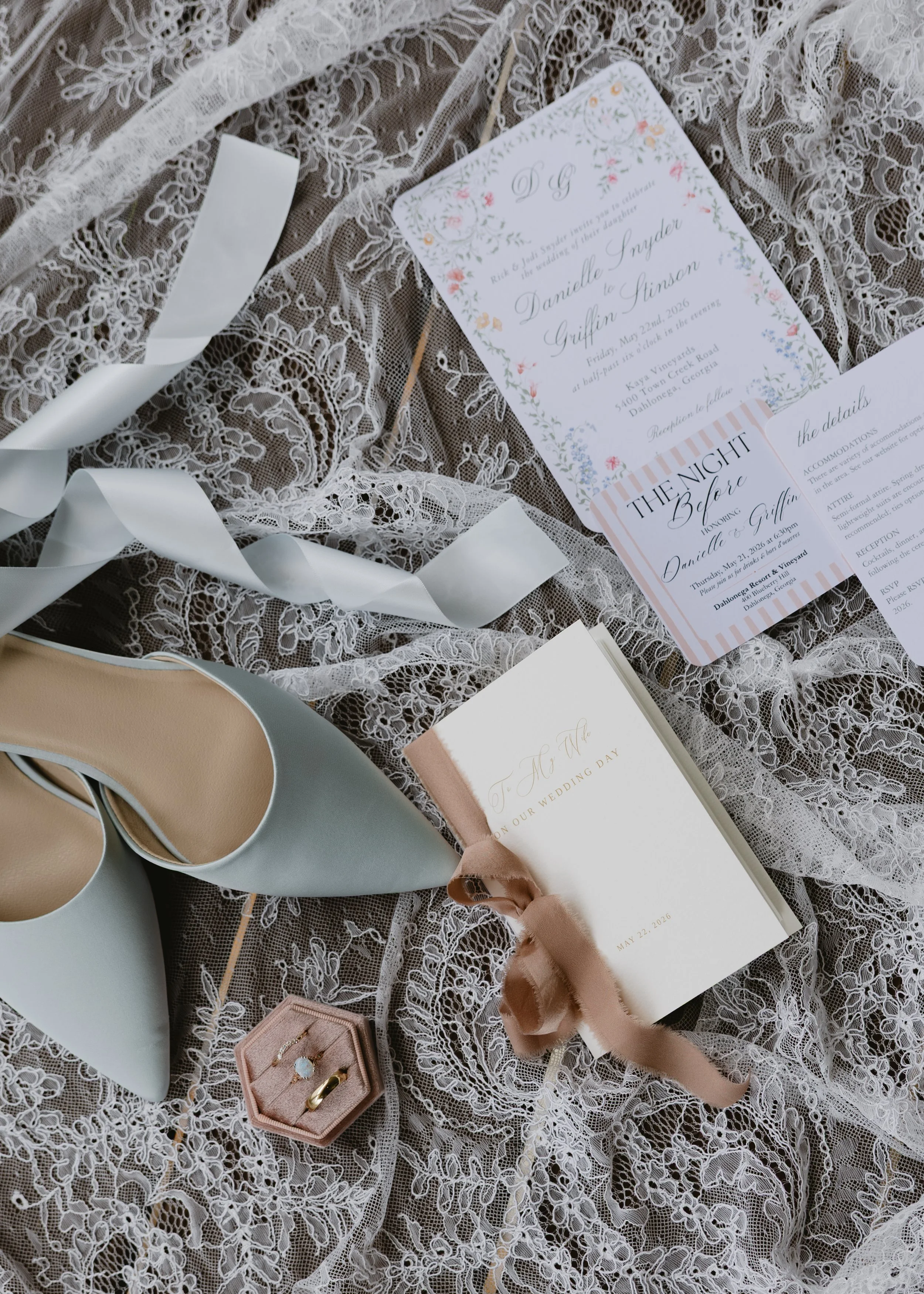 Wedding invitation, jewelry, a pair of white heels, and lace fabric on a table, ready for a wedding event.