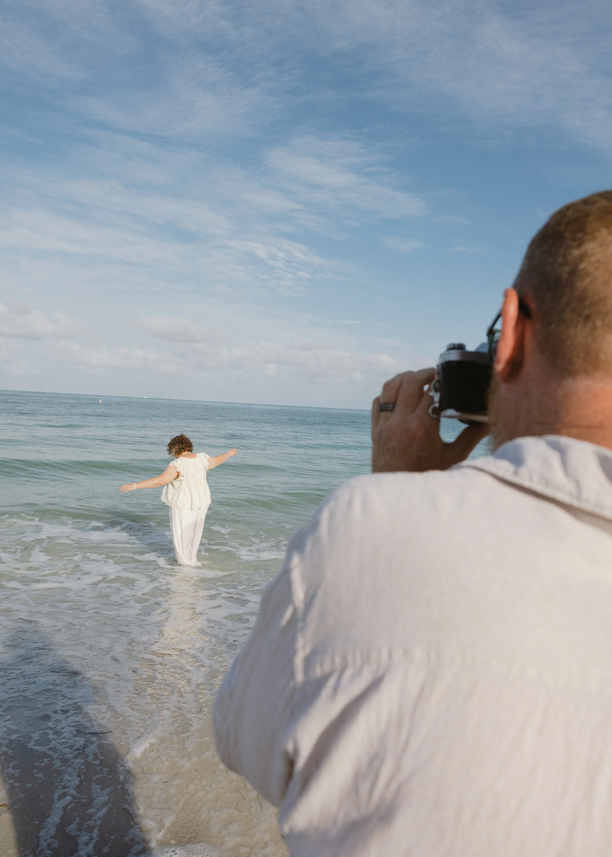 A person with blond hair is taking a picture of a woman standing in the ocean, wearing a white outfit with outstretched arms, near a beach with blue sky and clouds.