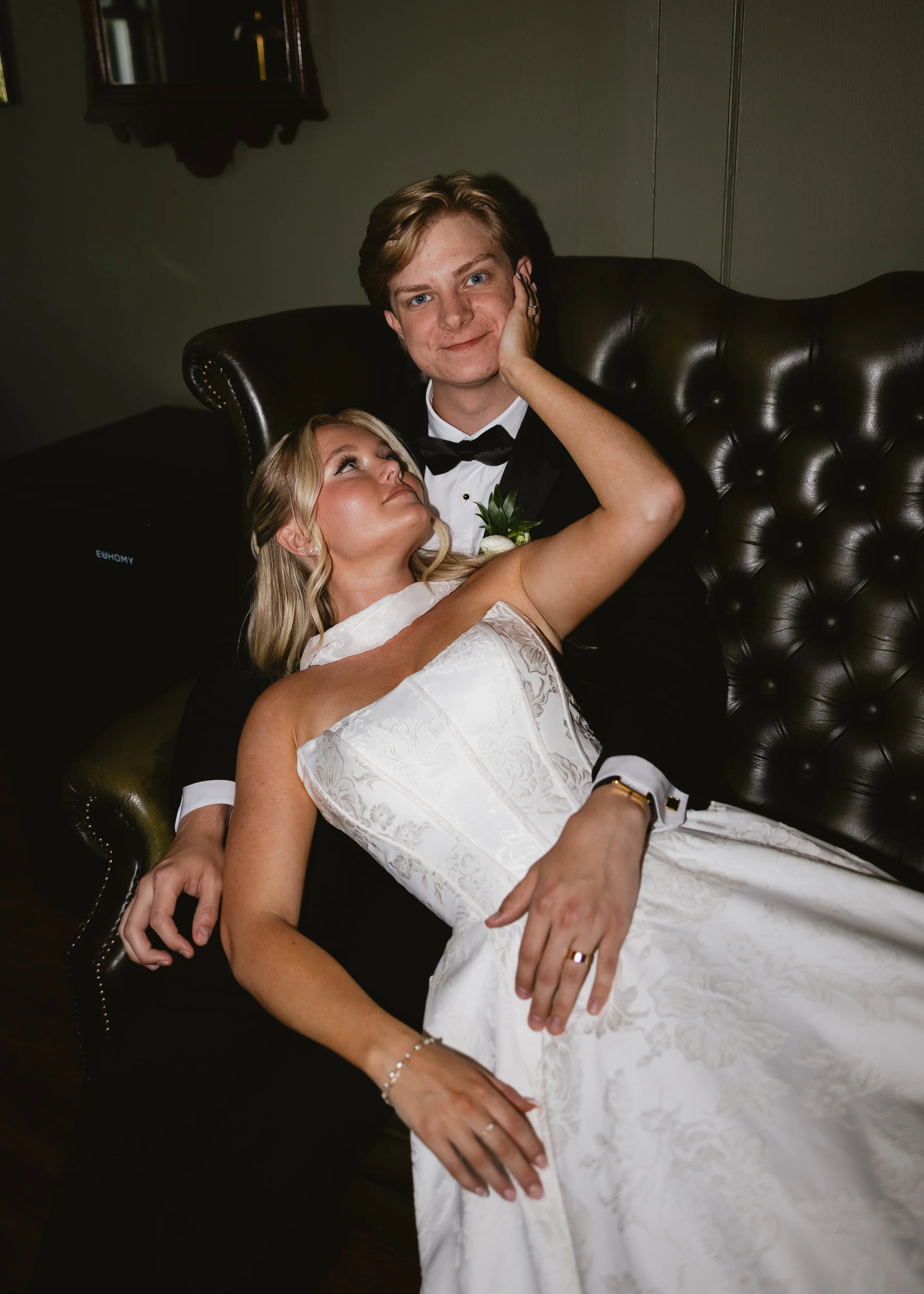 A bride and groom sitting on a dark leather sofa, with the bride in a white wedding dress and the groom in a black tuxedo with a bow tie, smiling and posing for a photo.