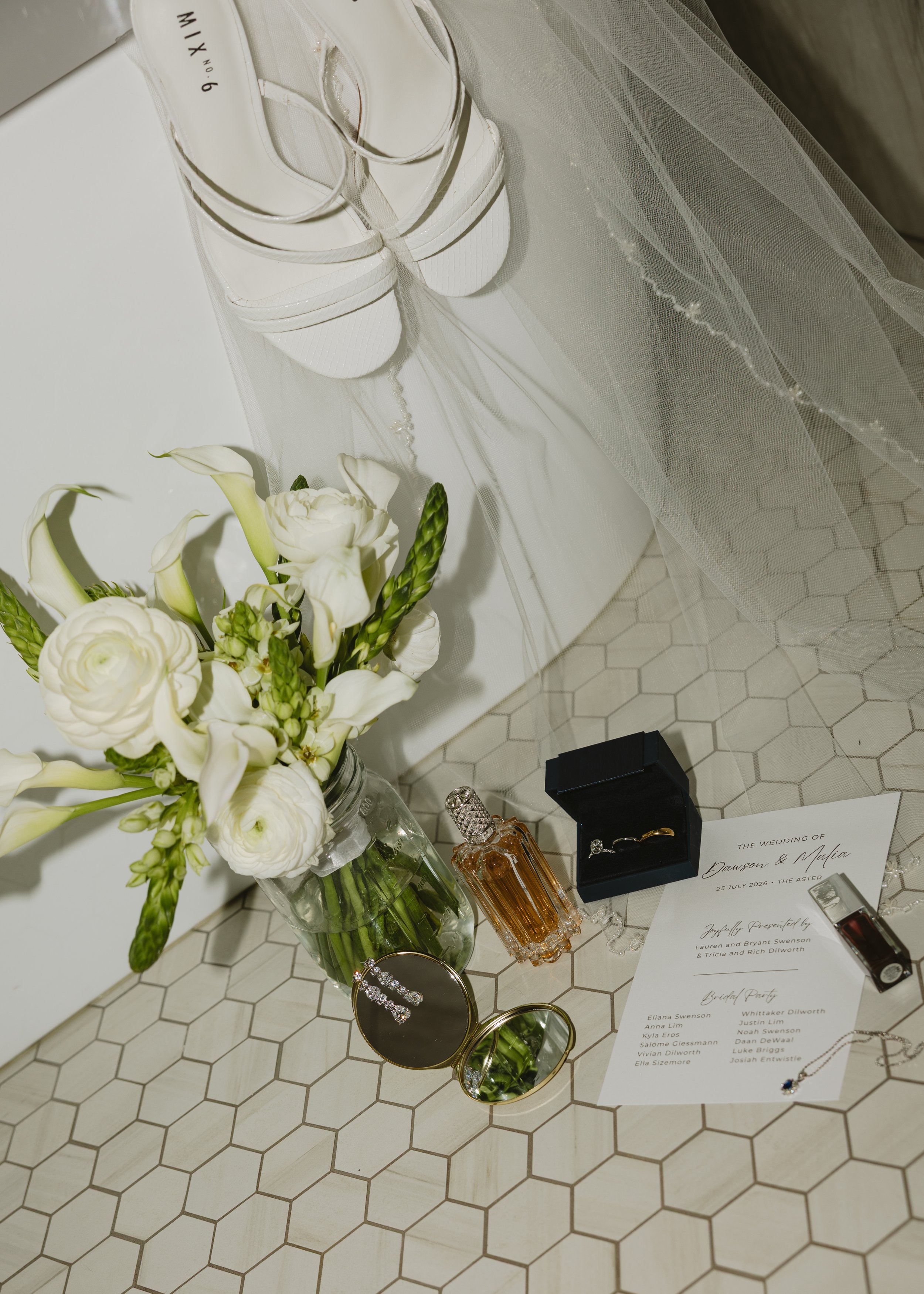 Wedding table display with white flowers in a glass jar, white sneakers, a jewelry box with rings, perfume bottles, and a wedding program on a hexagonal tiled floor.