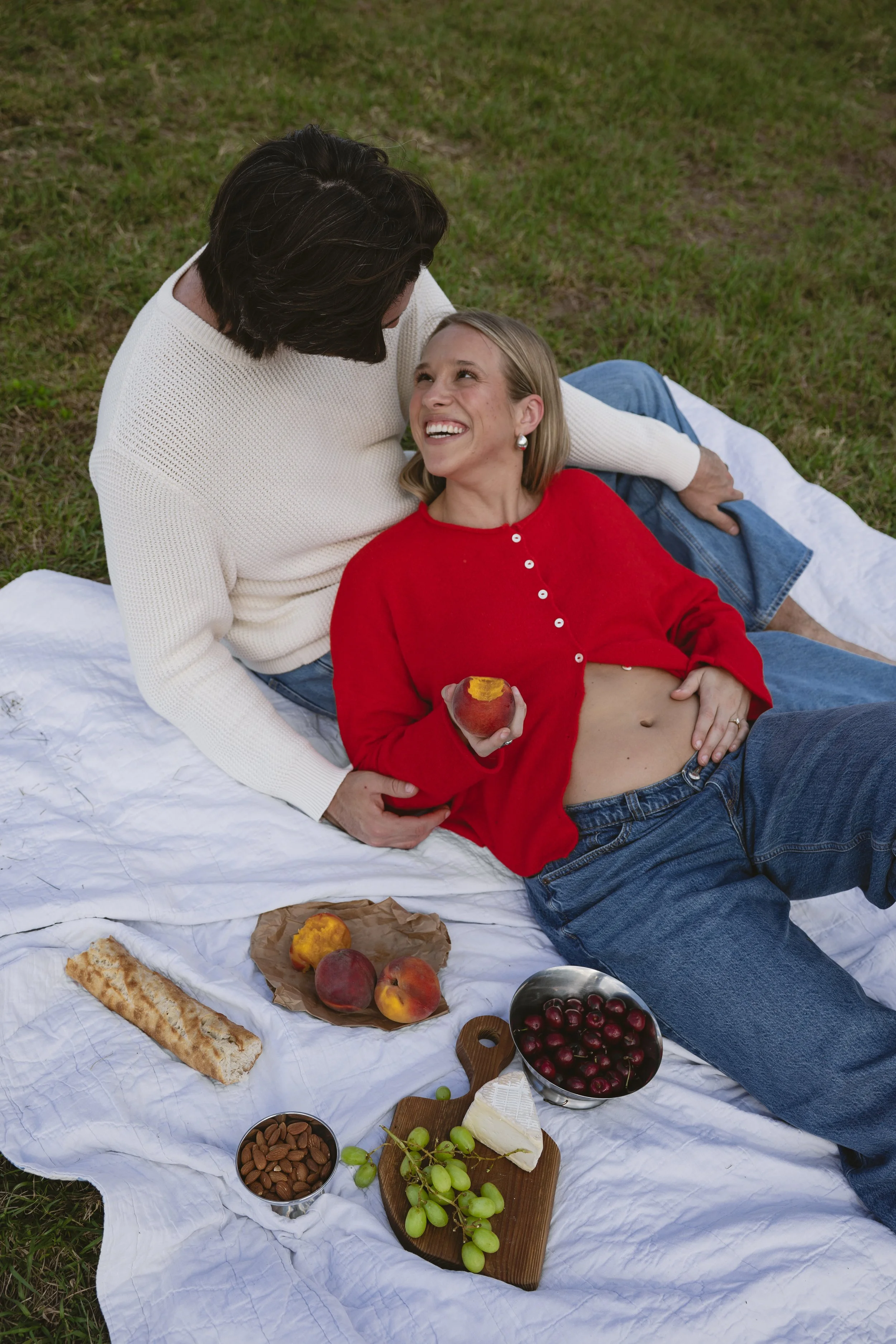 A woman lying on the grass with a man, enjoying a picnic. The woman is holding an apple and smiling at the man, who is looking at her. There is a white blanket on the grass with various foods including grapes, apples, a cheese wedge, almonds, a bread