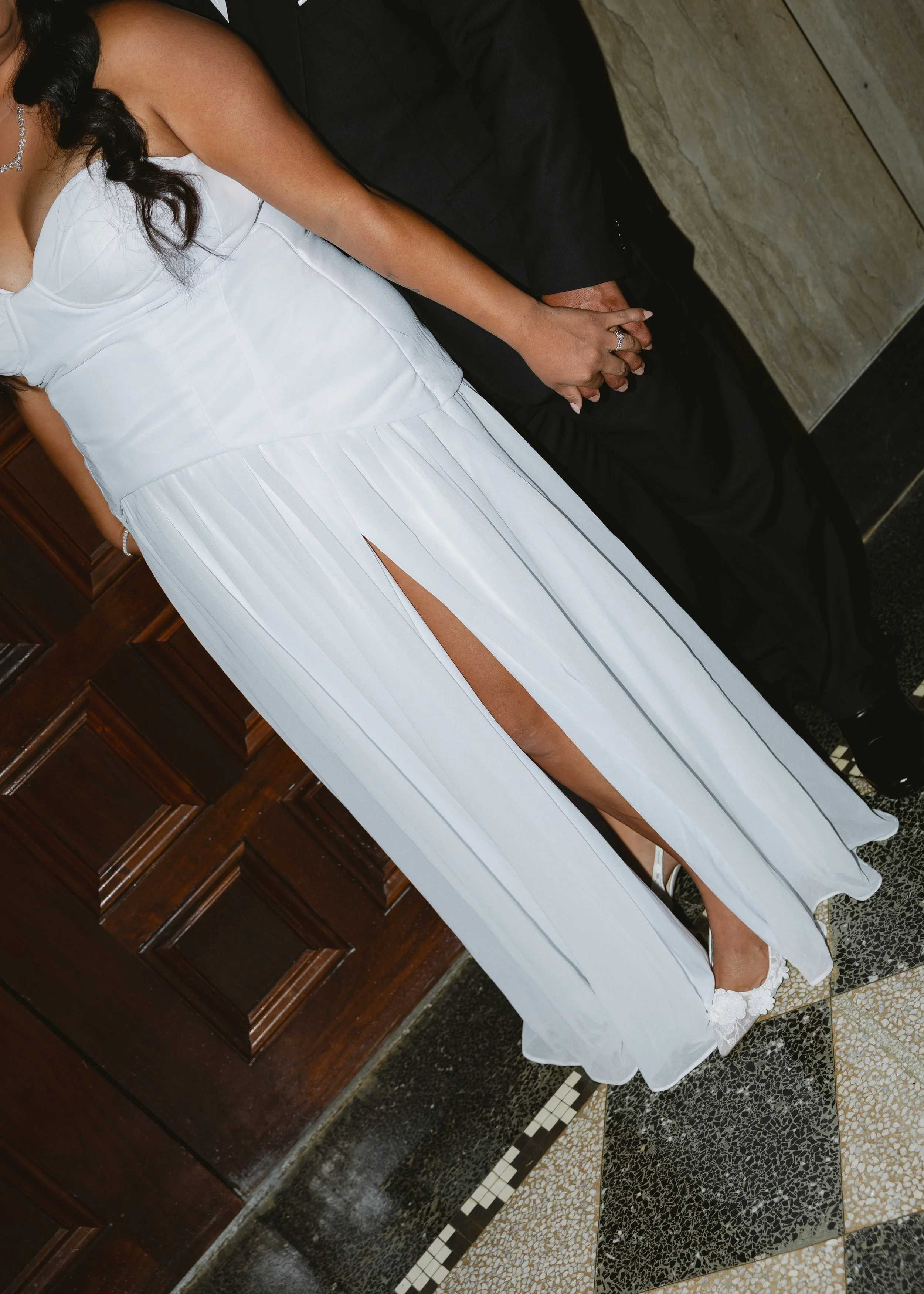 A woman in a white dress with a high slit, sitting on a stone bench, holding hands with a man in a black suit. The woman is wearing a silver necklace and white shoes.
