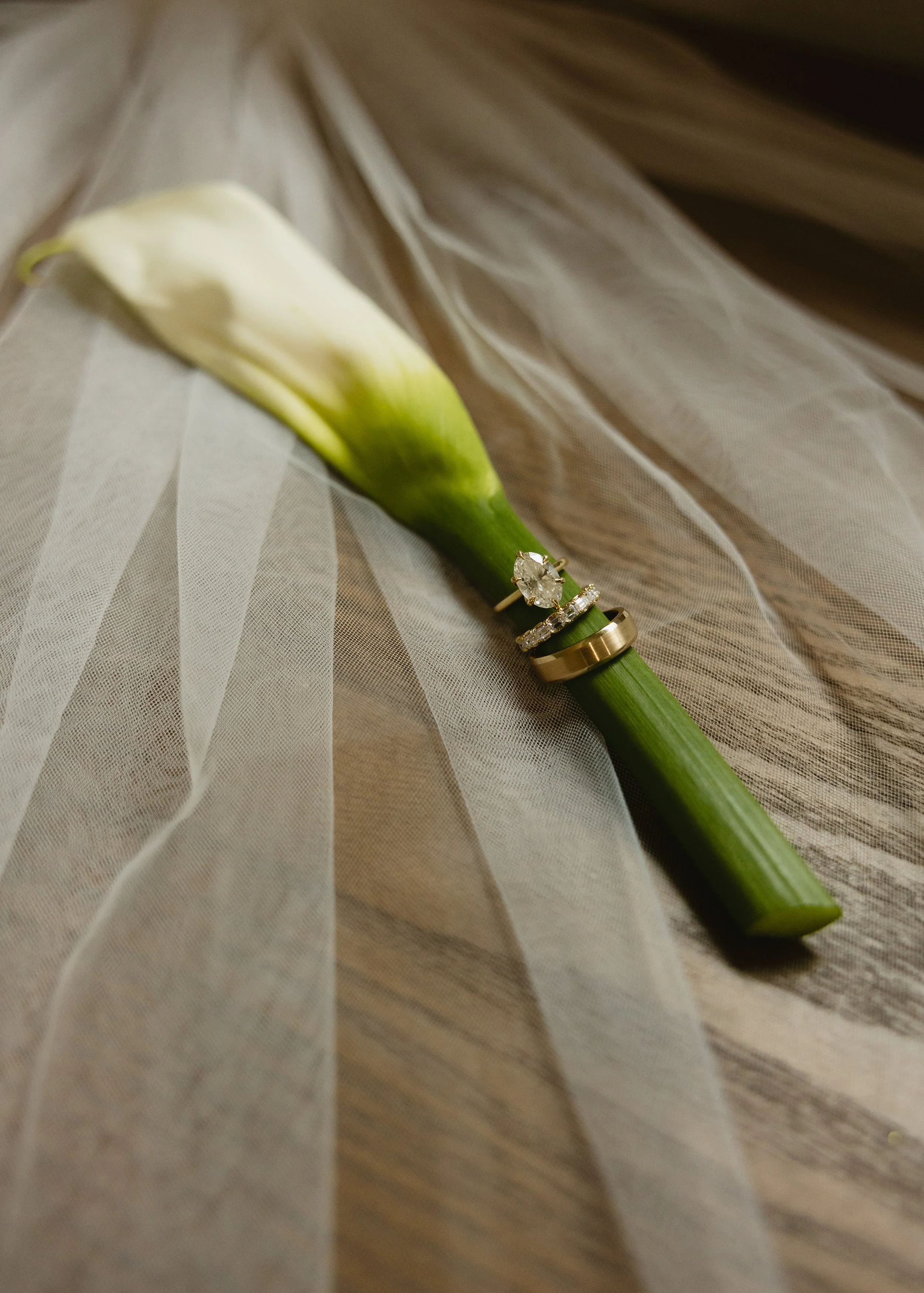 A white lily flower with wedding rings resting on its stem, placed on a sheer, flowing fabric on a wooden surface.