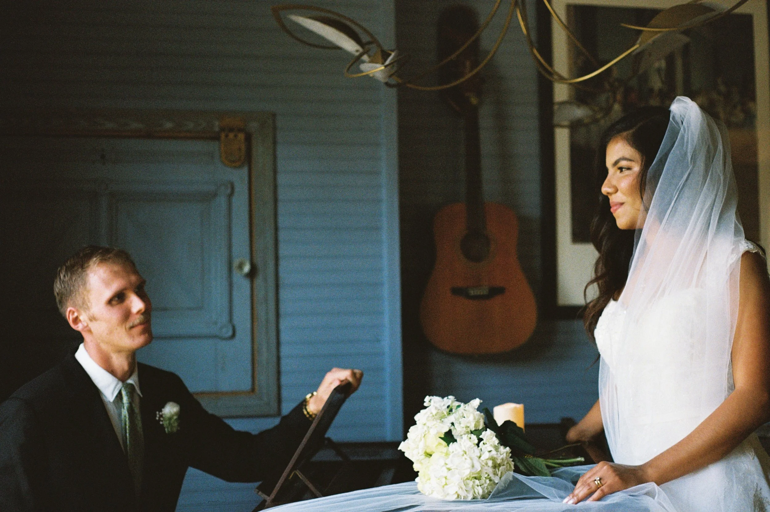 A bride dressed in white with a veil and a bouquet of flowers is at her wedding, exchanging rings with the groom, who is wearing a black suit.