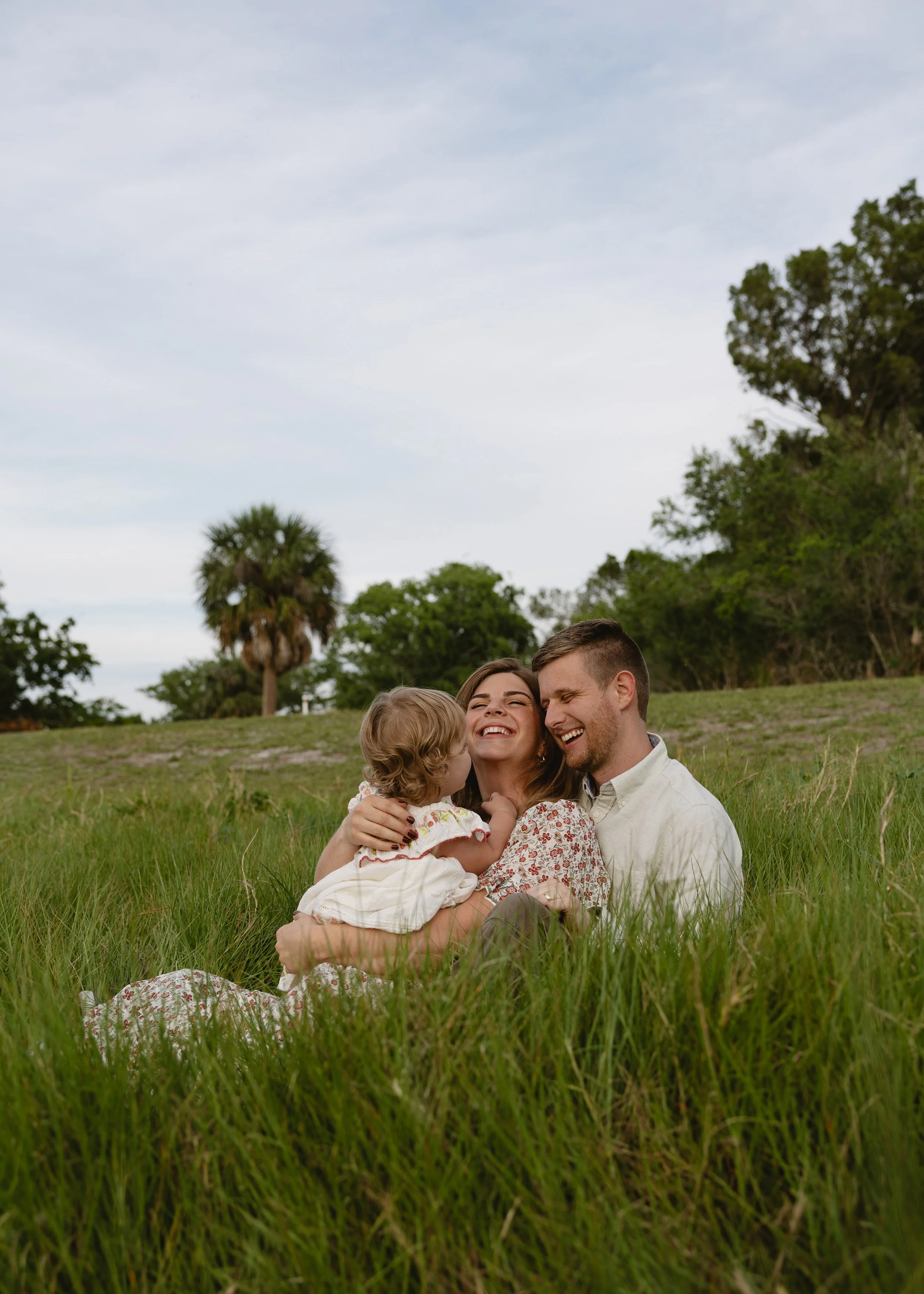 A happy family sitting on green grass outdoors, with trees and a cloudy sky in the background. The family includes a man, a woman, and a young girl, all smiling and enjoying each other's company.