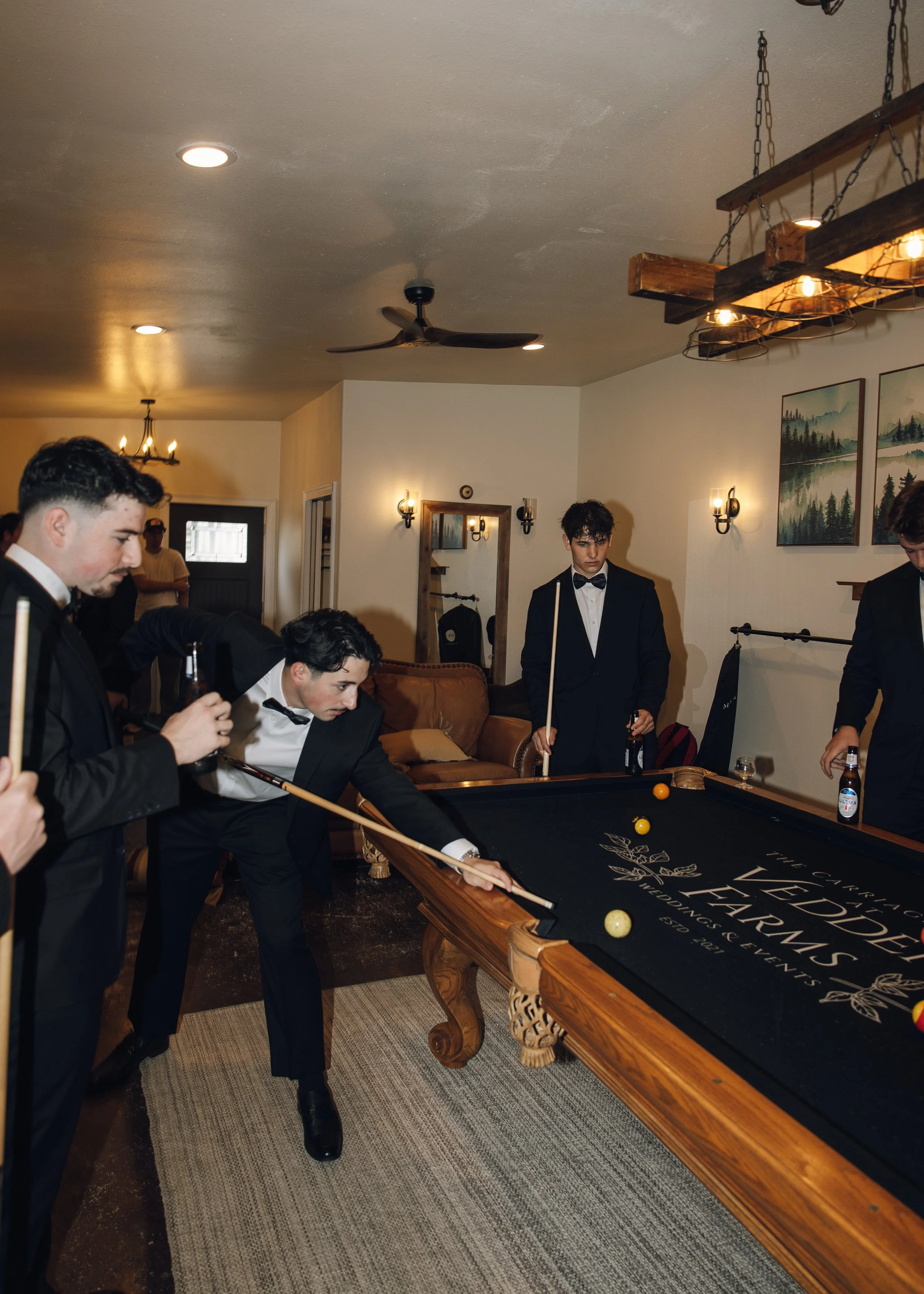 Group of young men dressed in tuxedos playing pool in a cozy, warmly lit room with rustic decor, including wall art, wood beams, and chandeliers.