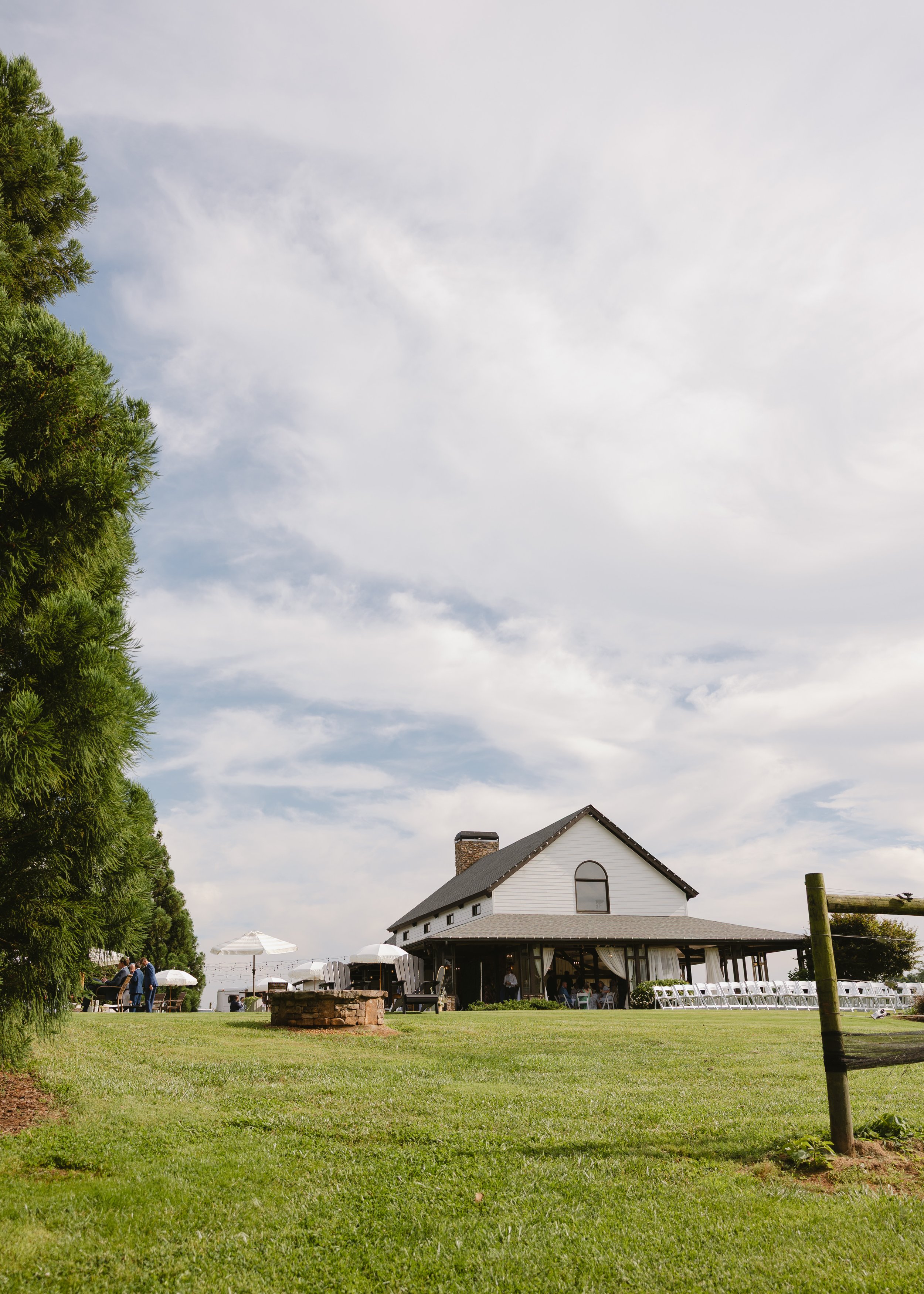 A large white house with a gabled roof and a chimney, set in a grassy area with outdoor seating, umbrellas, and a few people enjoying a gathering. There are trees on the left and a cloudy sky overhead.