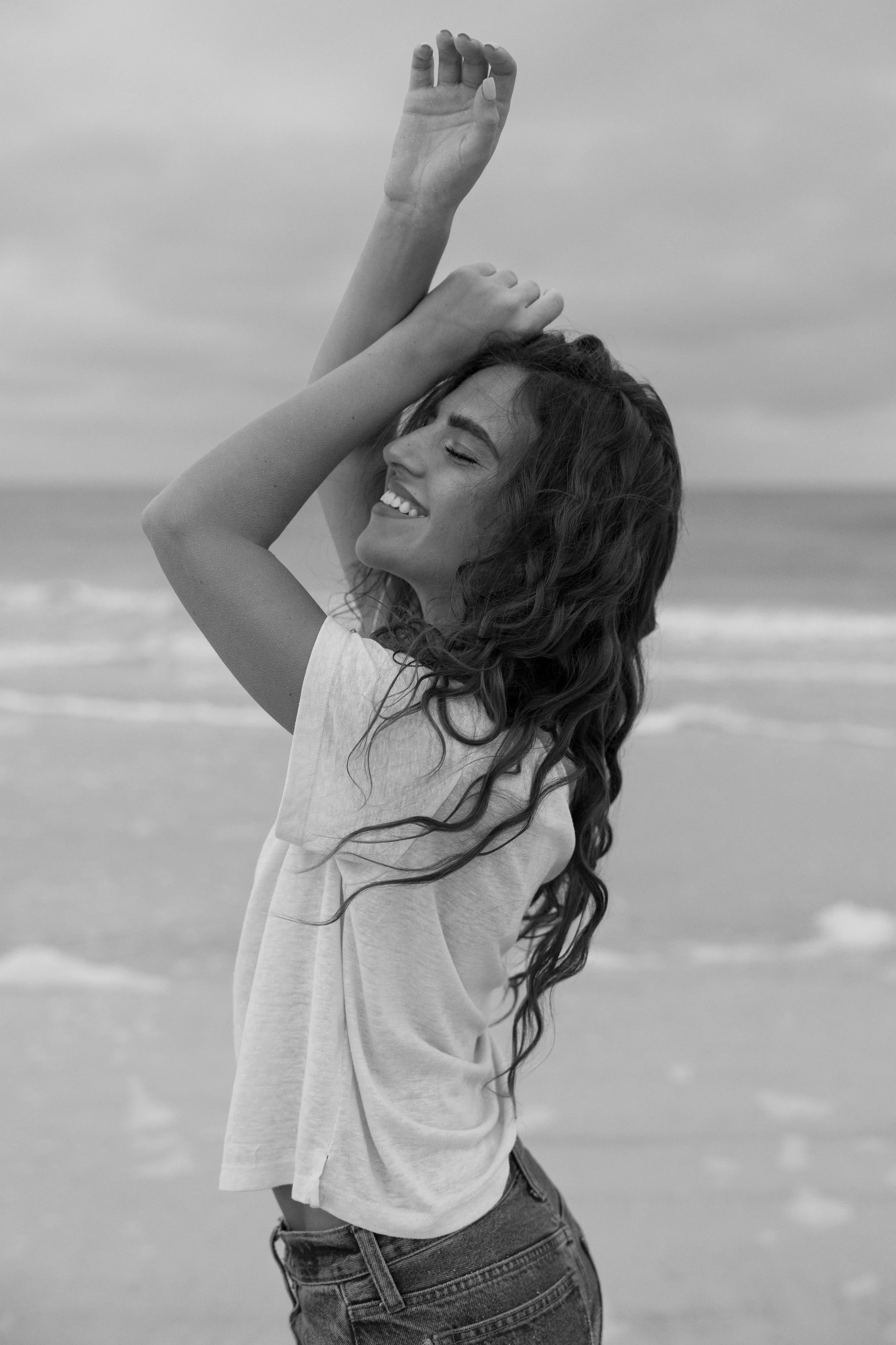 A woman with long, curly hair smiling with eyes closed on a beach, wearing a loose t-shirt and jeans, with waves in the background.