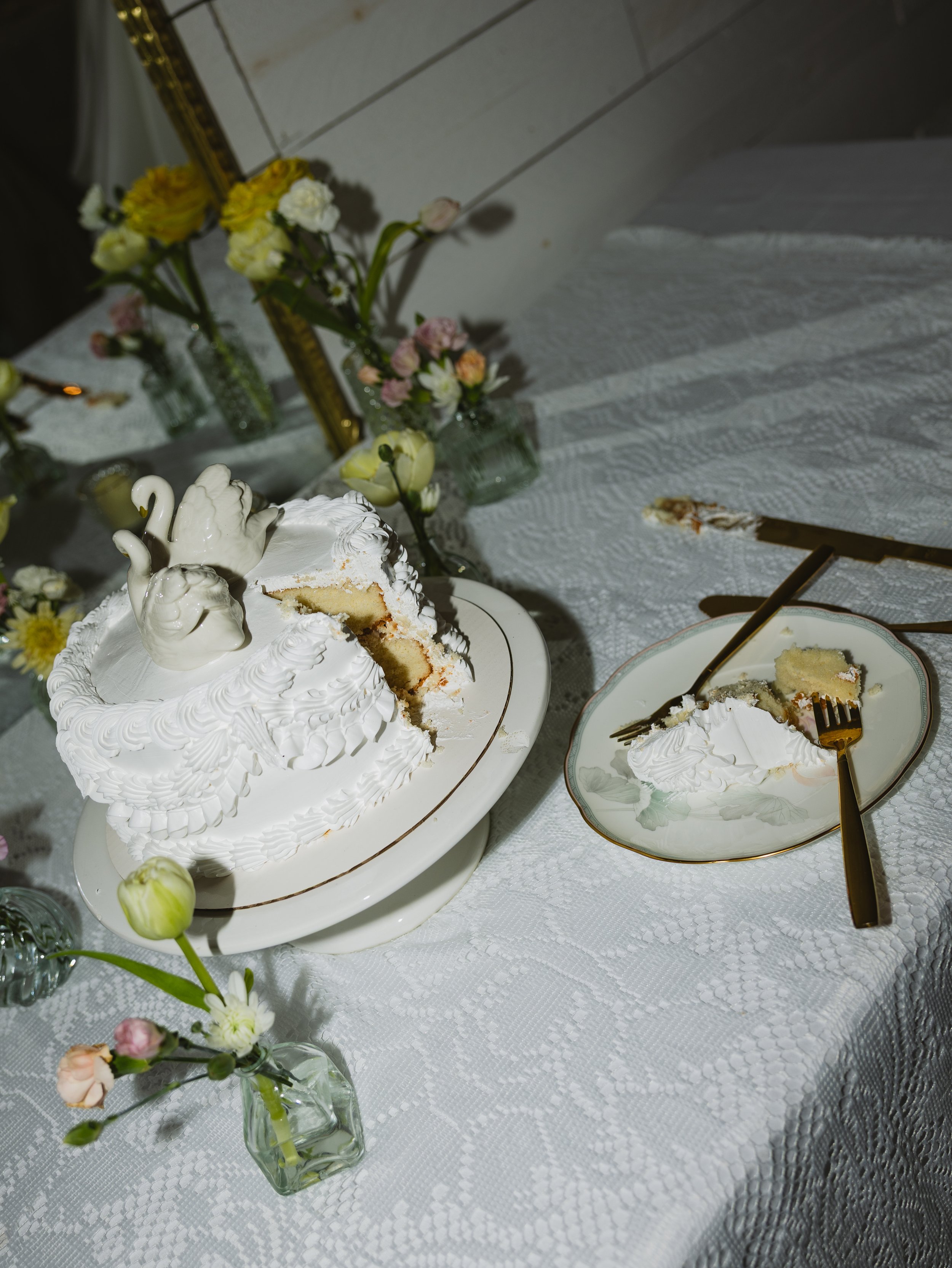 A white cake decorated with swan-shaped figures and piped icing, served on a white plate, with a slice taken out. The table has a white patterned tablecloth, with small vases of colorful flowers and bottles around.
