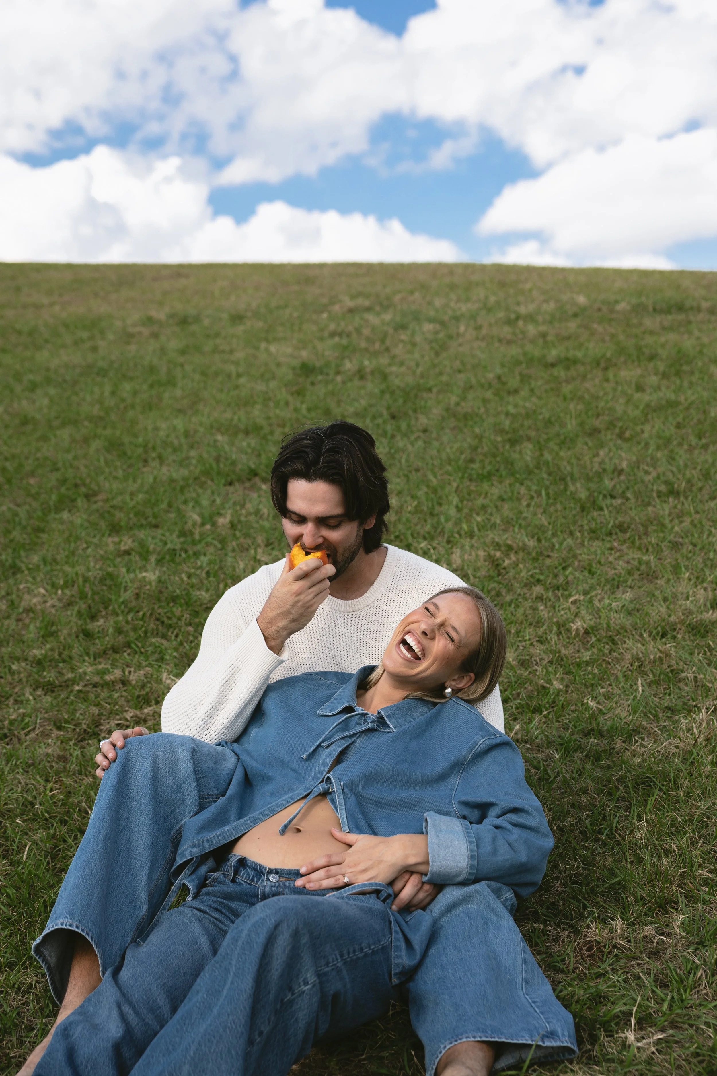 A couple lying on the grass, the man is eating a peach while the woman laughs and leans back.