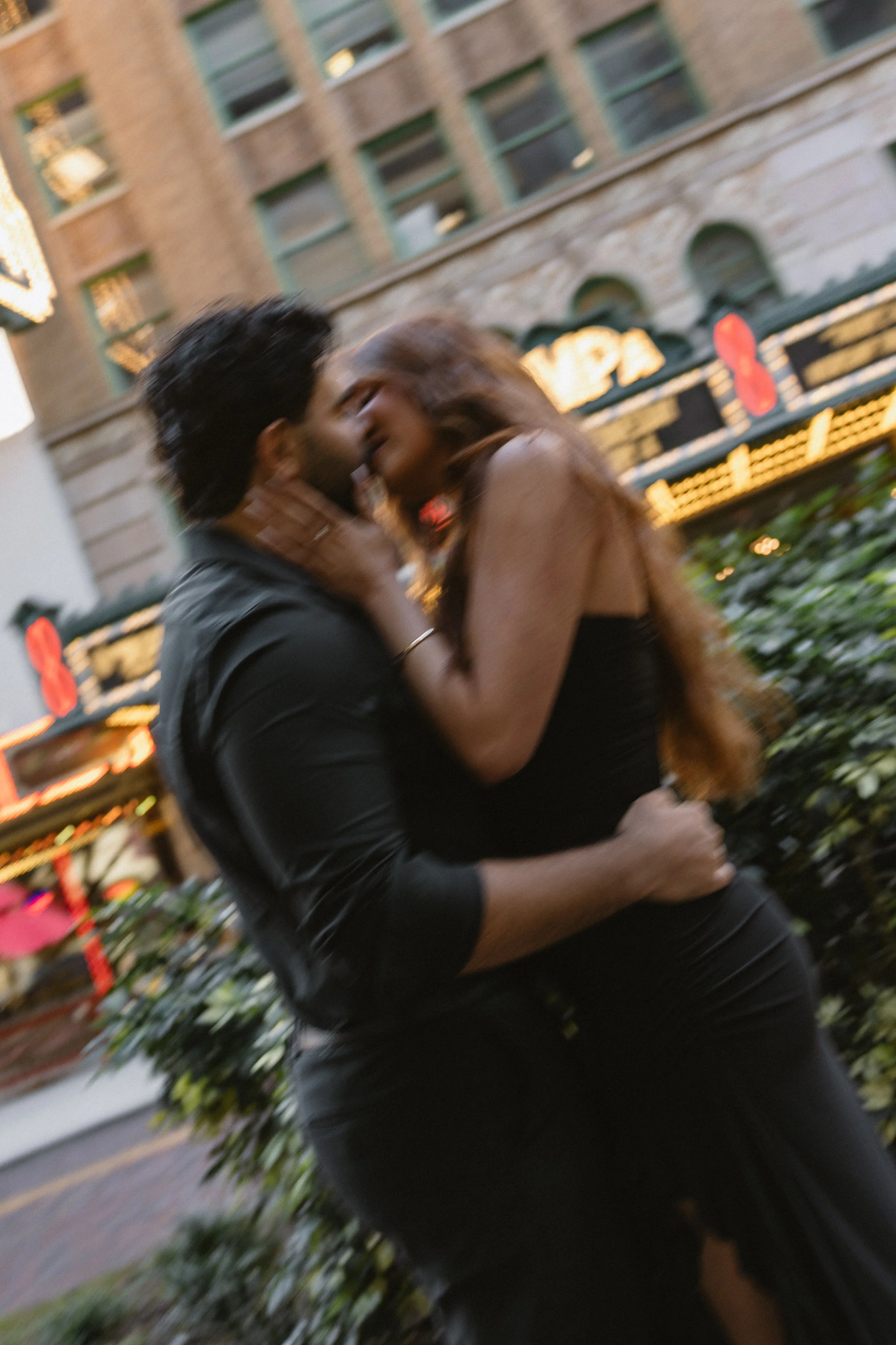 A couple sharing a kiss outdoors on a city street at night, with buildings and illuminated signs in the background.