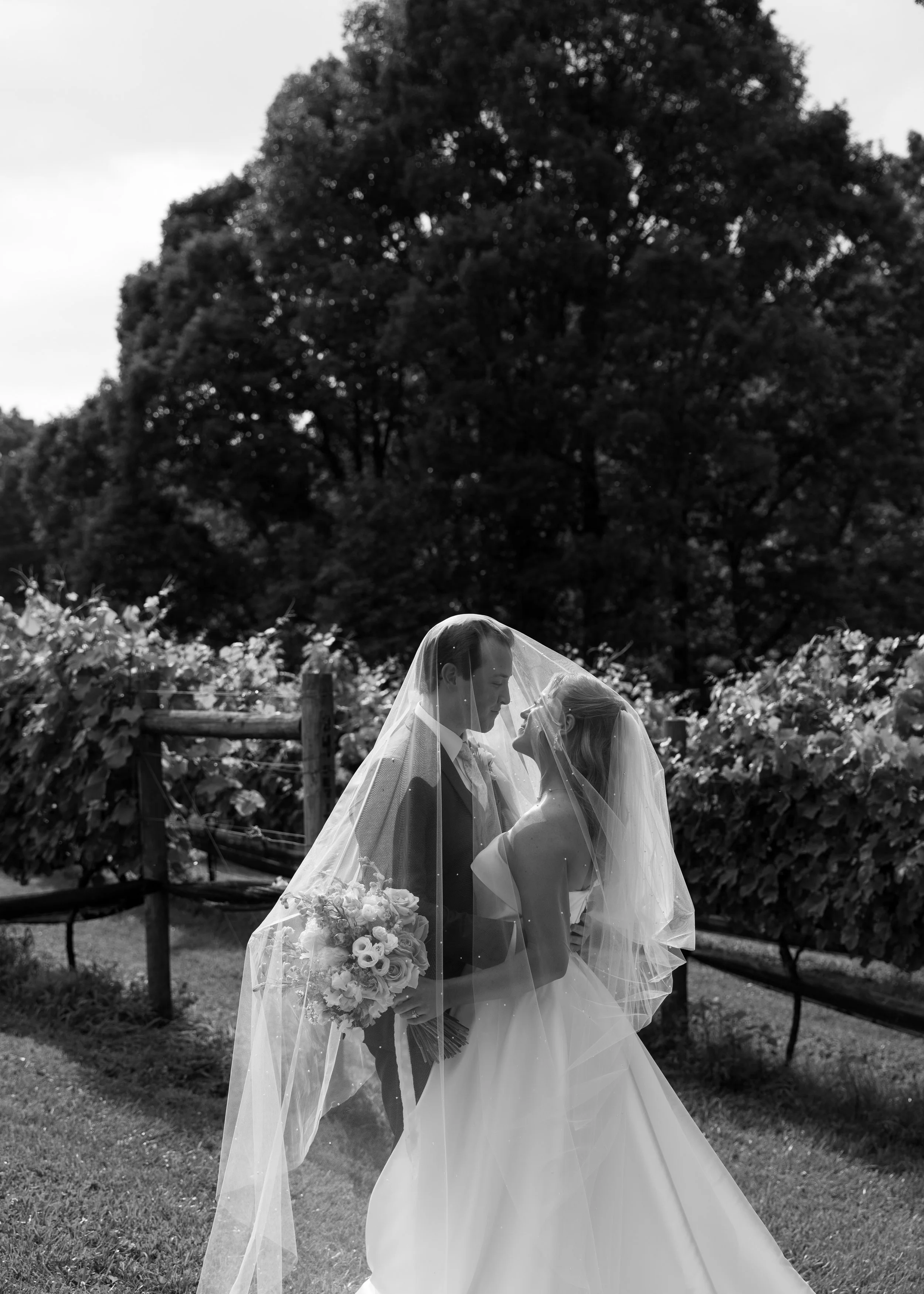 A bride and groom standing close together outdoors under a wedding veil, with trees and a fence in the background, in black and white.