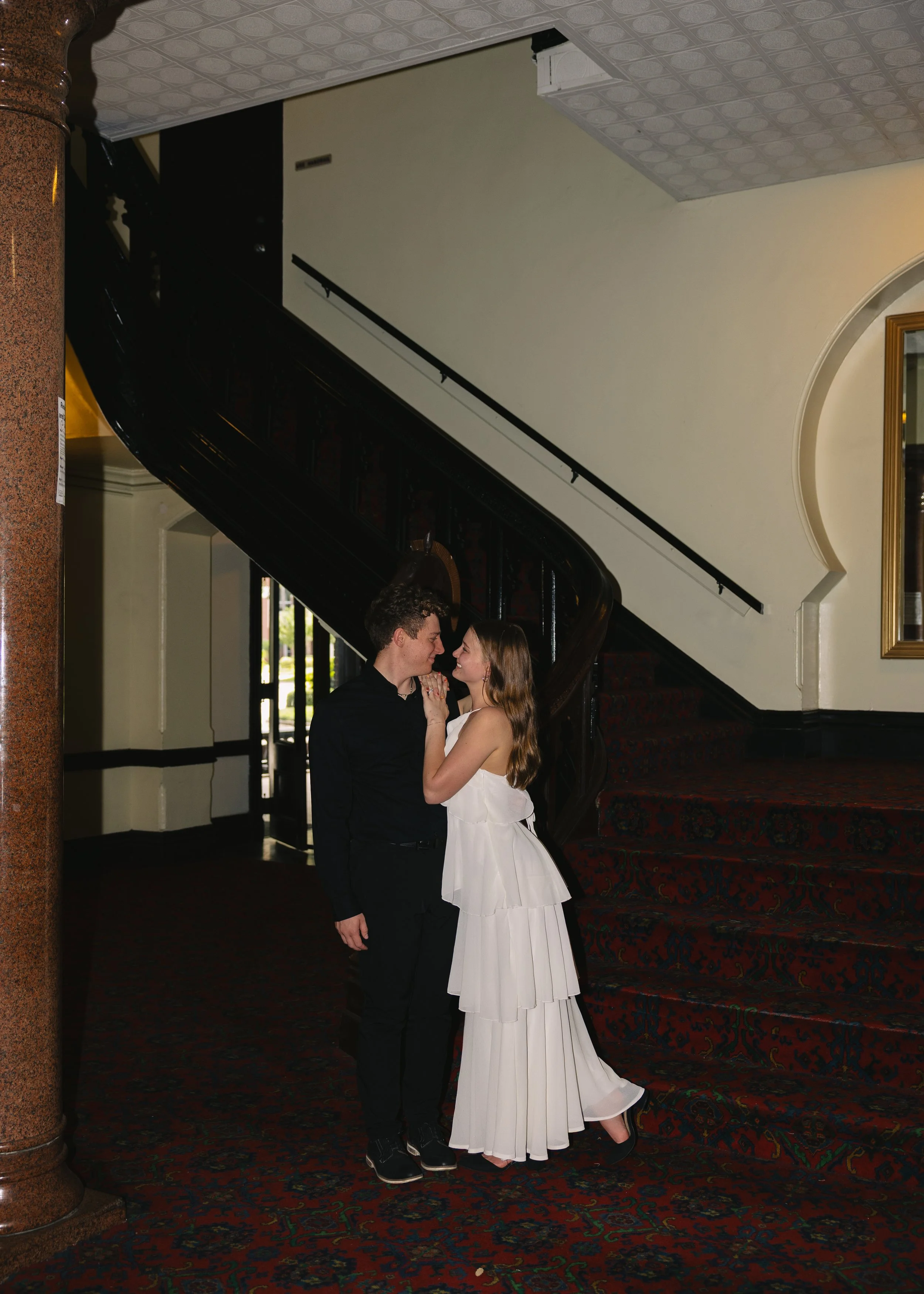A couple, dressed in formal attire, is standing close together near a staircase inside a building. The man is wearing a black suit, and the woman is in a white dress. They are looking into each other's eyes and holding hands.