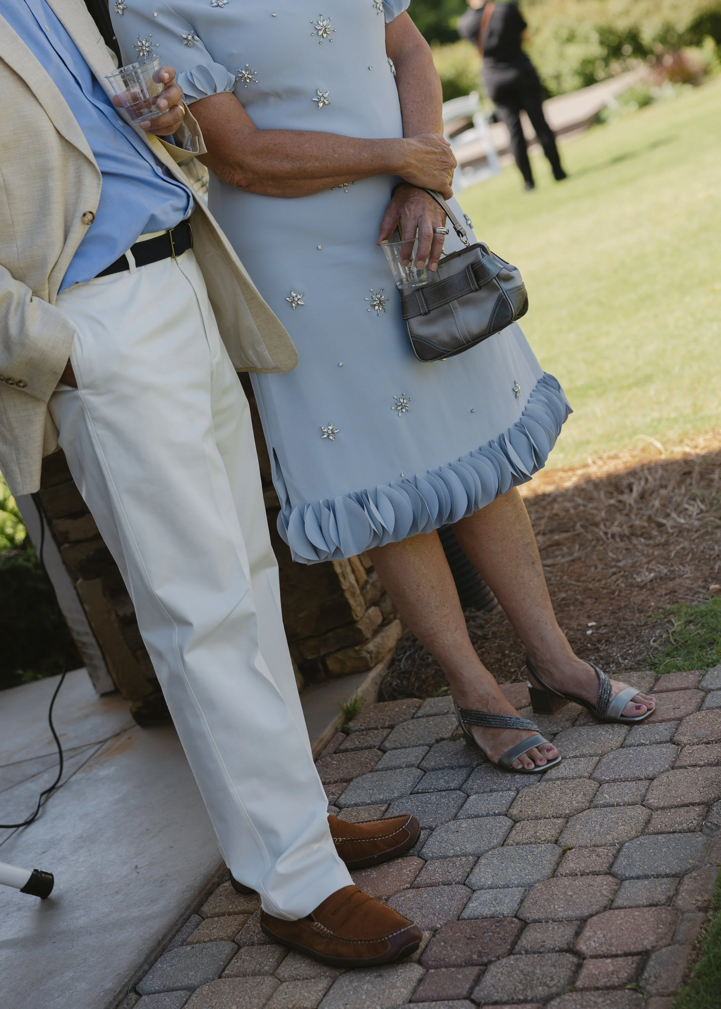 Close-up of pedestrians at an outdoor gathering, showing women in dresses and a man in a suit, all holding drinks, with greenery and a pathway in the background.