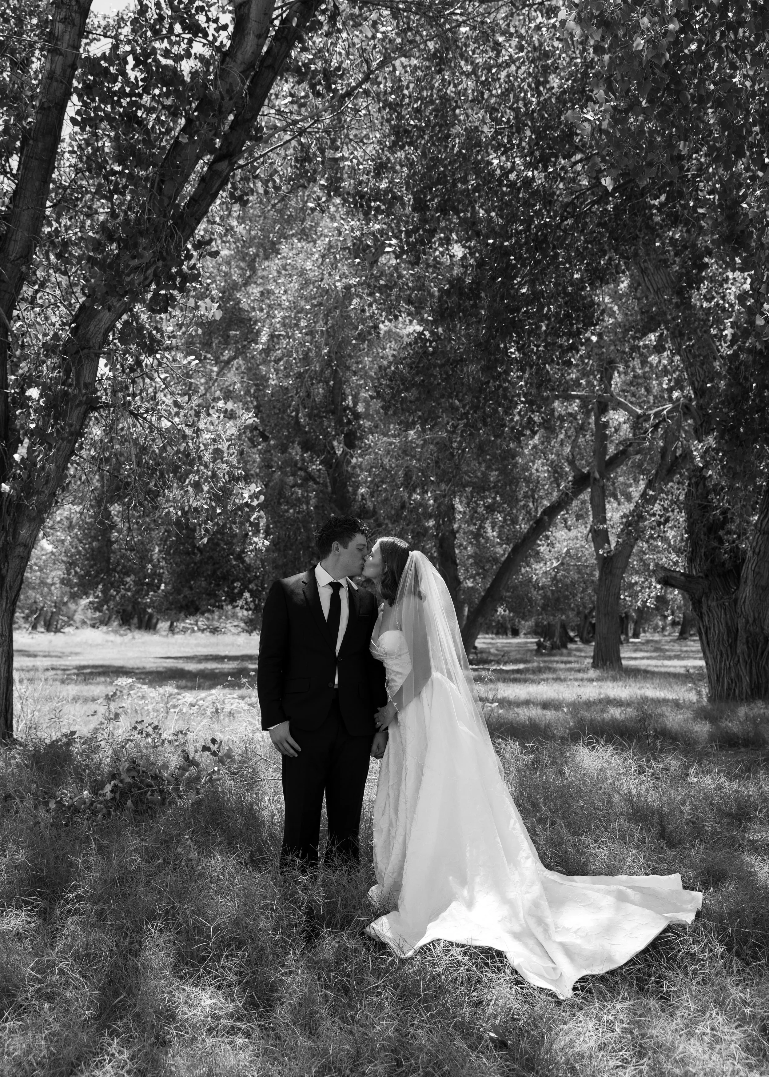 A black and white photo of a bride and groom standing close together in a wooded area, sharing a kiss. The bride is wearing a long white wedding gown with a veil, and the groom is dressed in a dark suit with a tie.