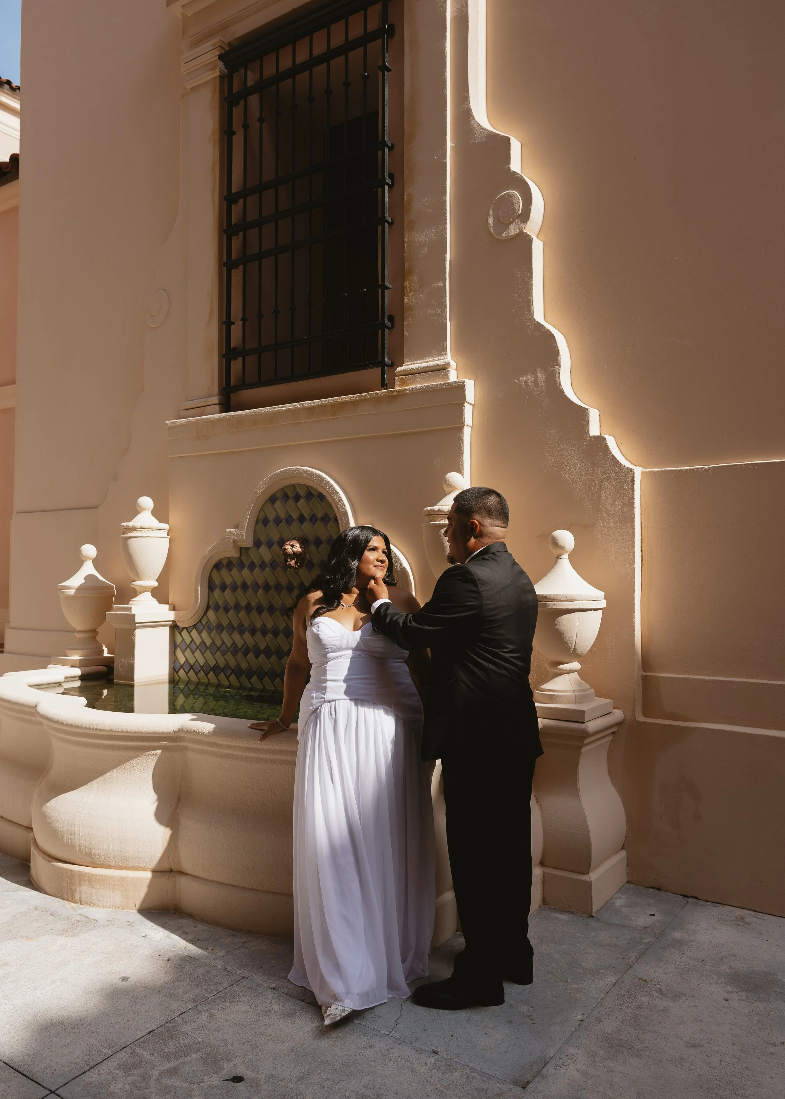 A couple stands by a decorative stone fountain in front of a beige stucco wall with architectural details and a window with black iron bars. The woman is wearing a white strapless dress, and the man is in a black suit. They are facing each other, eng