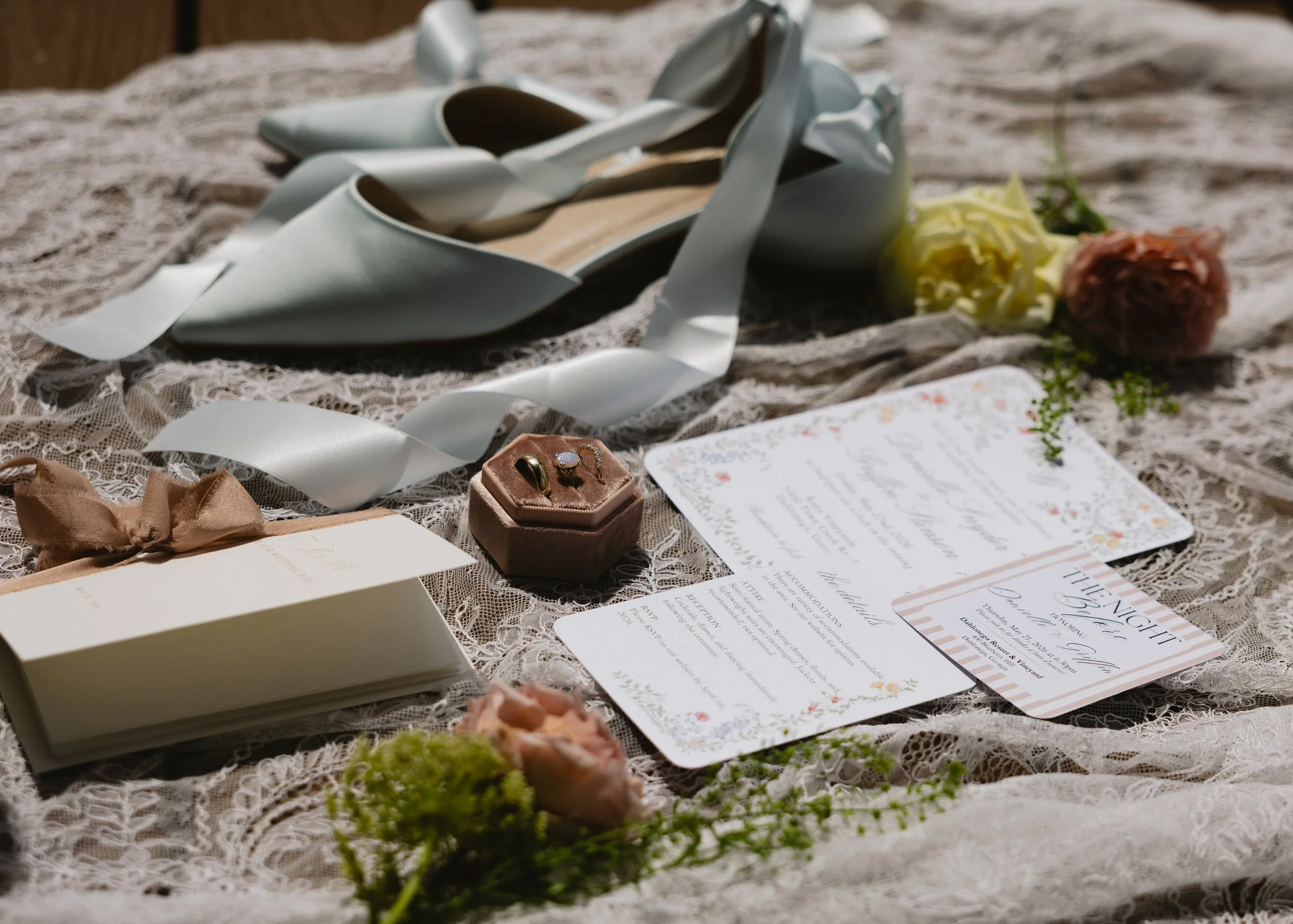 A lace tablecloth with wedding or event invitations, flowers, jewelry in a small box, and a pair of high heels.