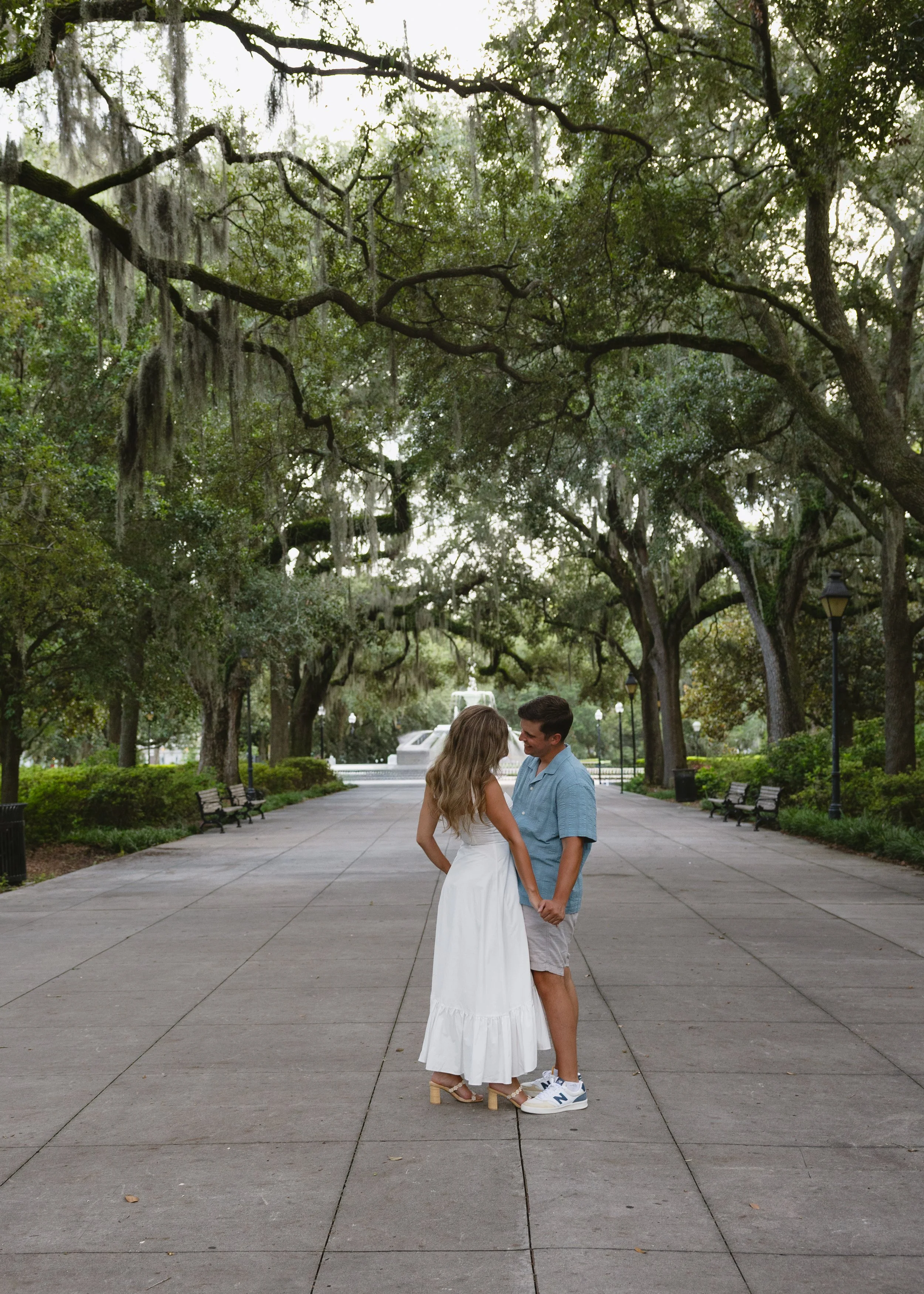 A young couple holding hands and looking at each other in a park with large trees, benches, and a water fountain in the background.