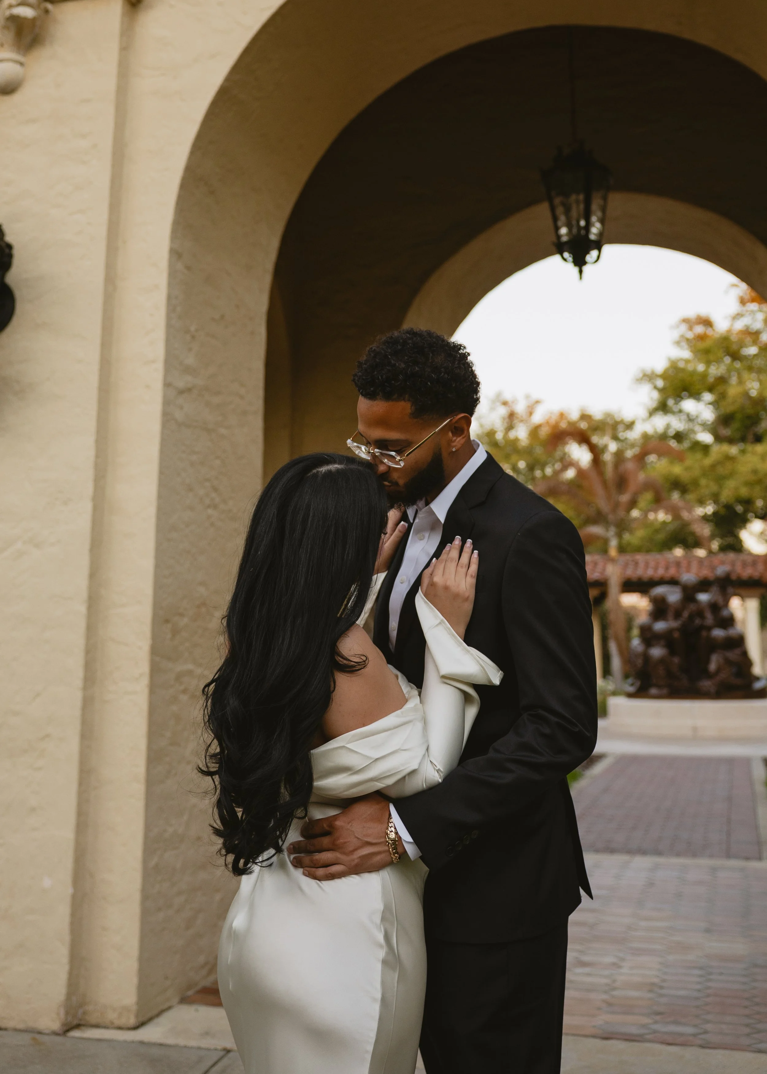 A couple dressed in formal attire sharing an intimate moment outdoors under an archway, with trees and a building in the background.