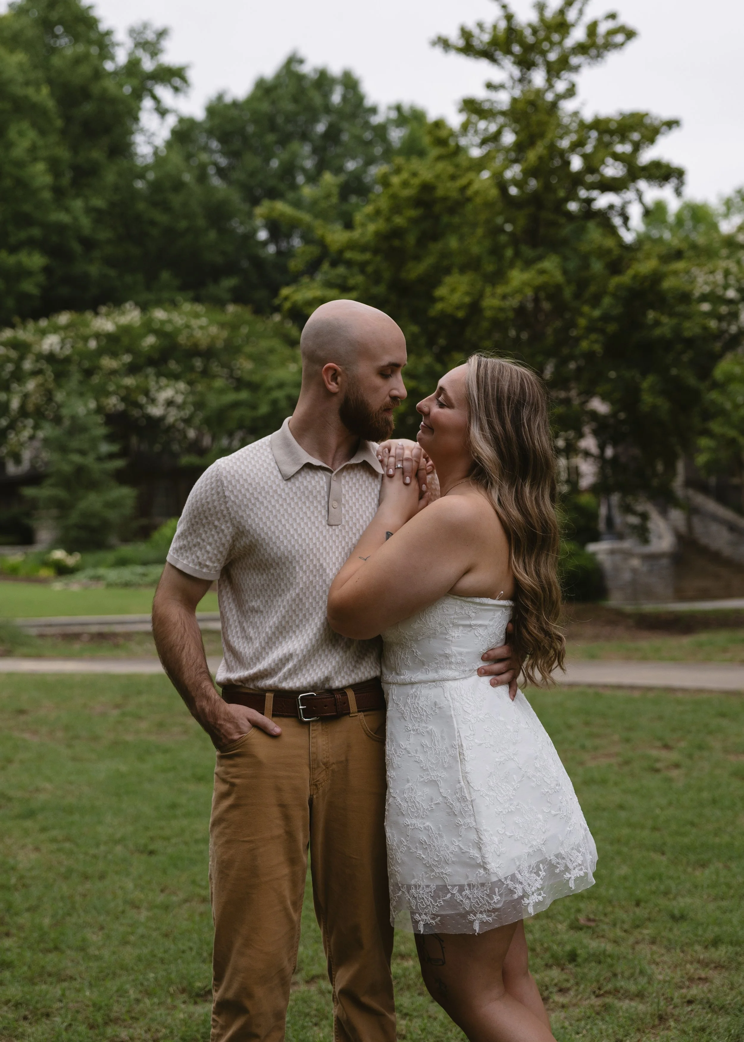 A man and a woman share a tender moment outdoors, standing close with their foreheads touching and eyes closed, in a park with green trees and a small stream in the background.