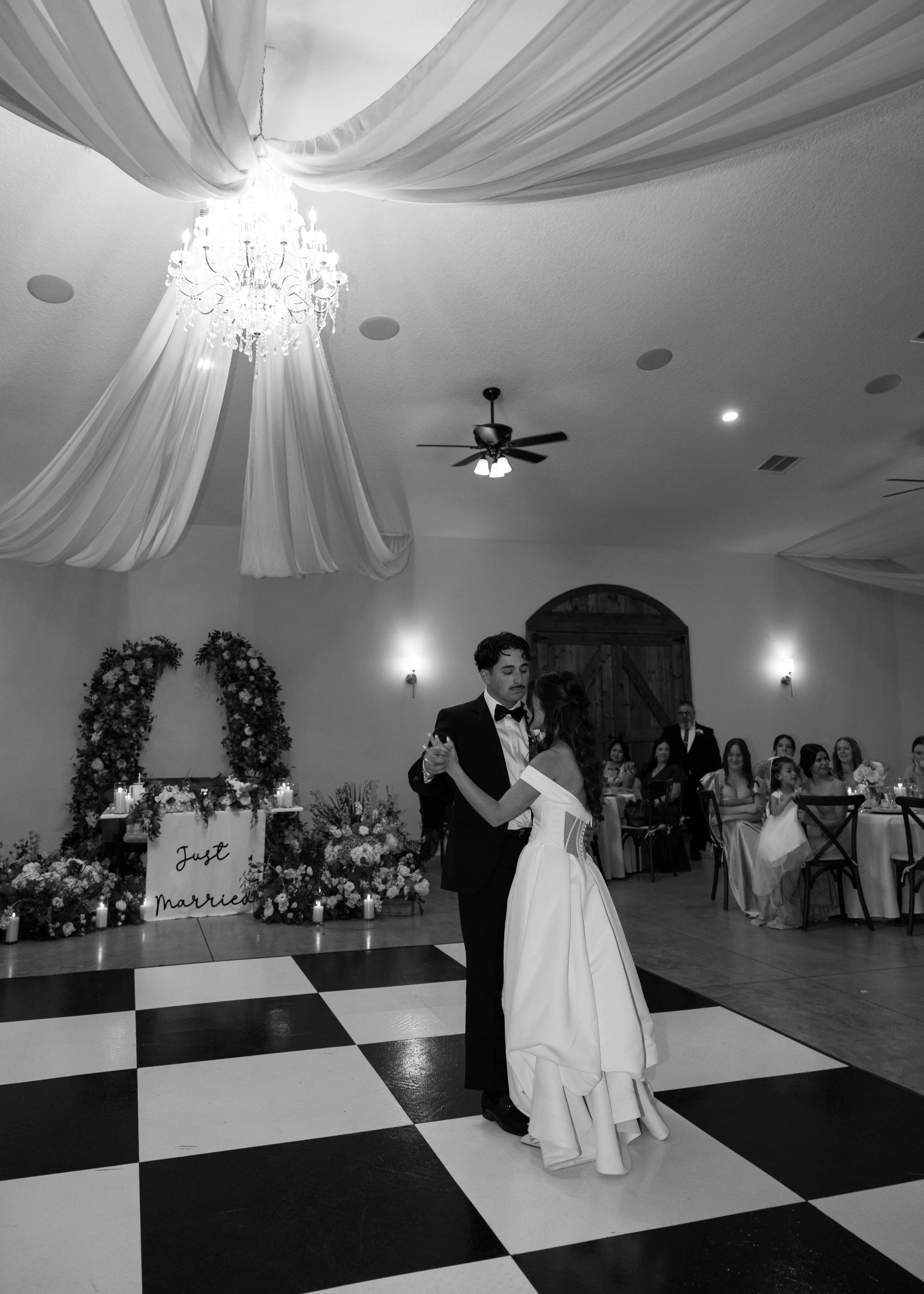 A couple is dancing at their wedding reception. The groom is wearing a black tuxedo with a bow tie, and the bride is in a white wedding gown. They are dancing on a black and white checkered dance floor. In the background, guests are seated at tables 