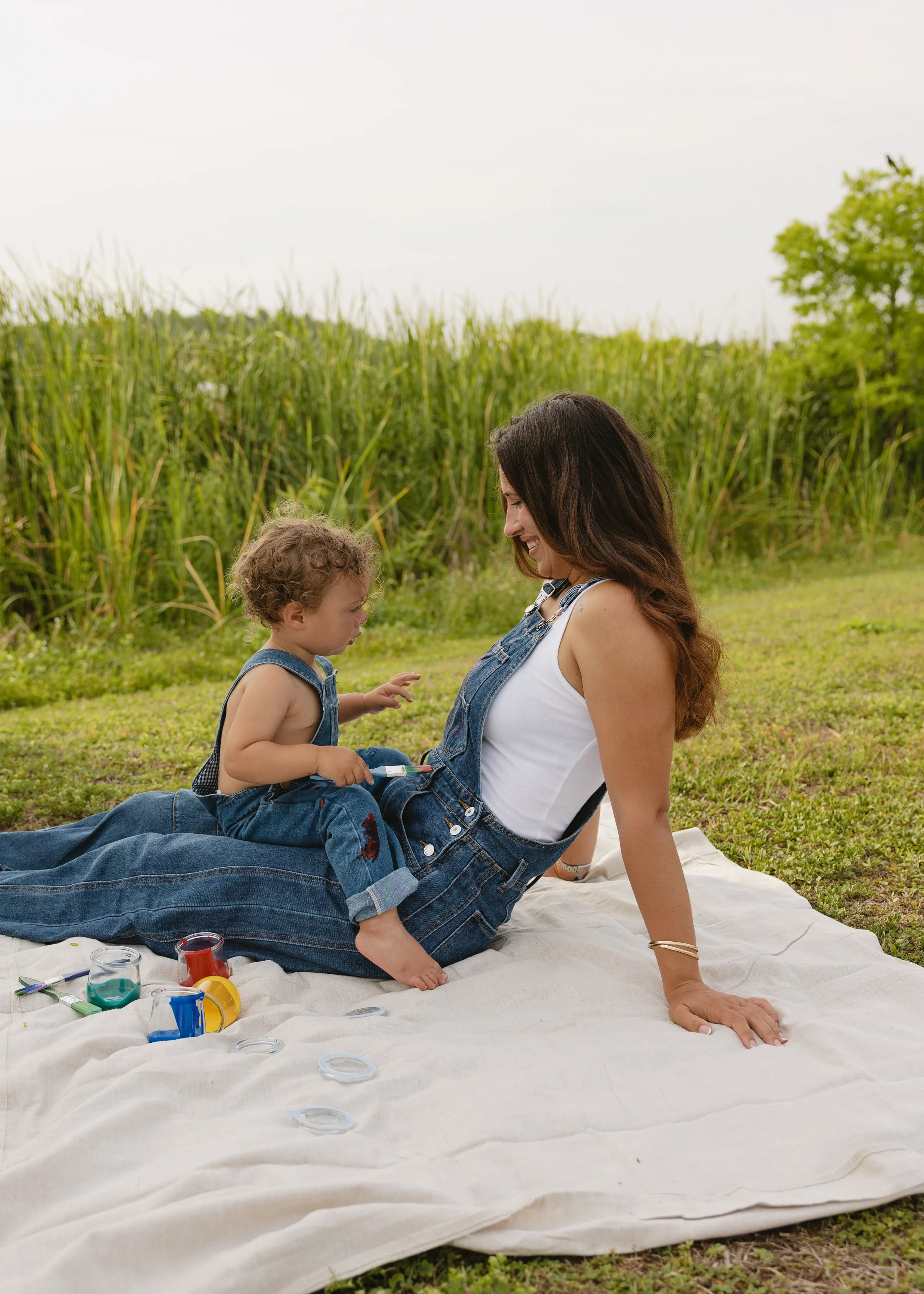A woman with long dark hair and a young boy sitting on a white blanket outdoors. The woman is smiling at the boy, who is looking down, and they are surrounded by jars of paint, brushes, and art supplies. The background features tall green grass and t