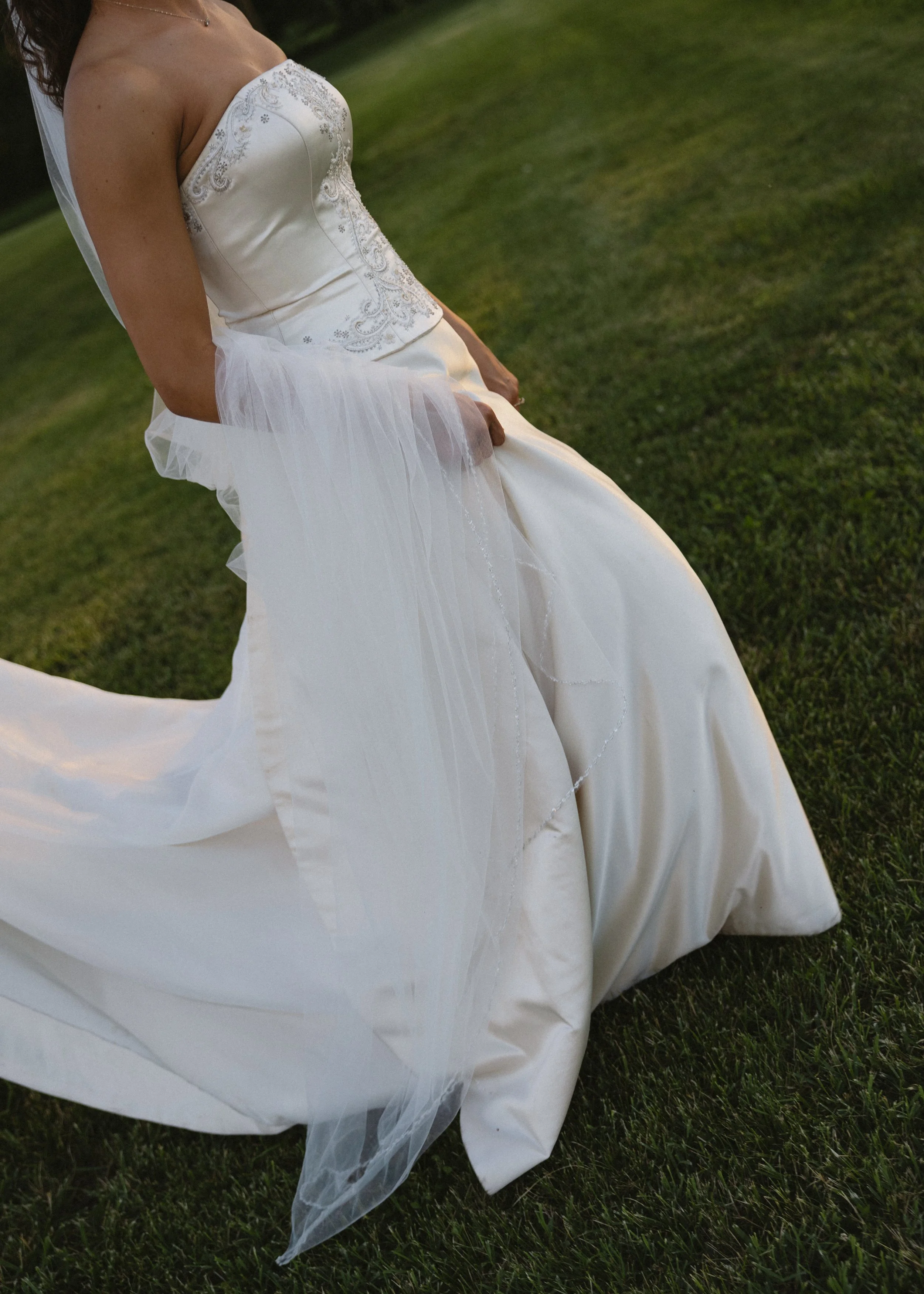 A bride in a white wedding dress holding the skirt of her dress with one hand, standing on grass during daylight.