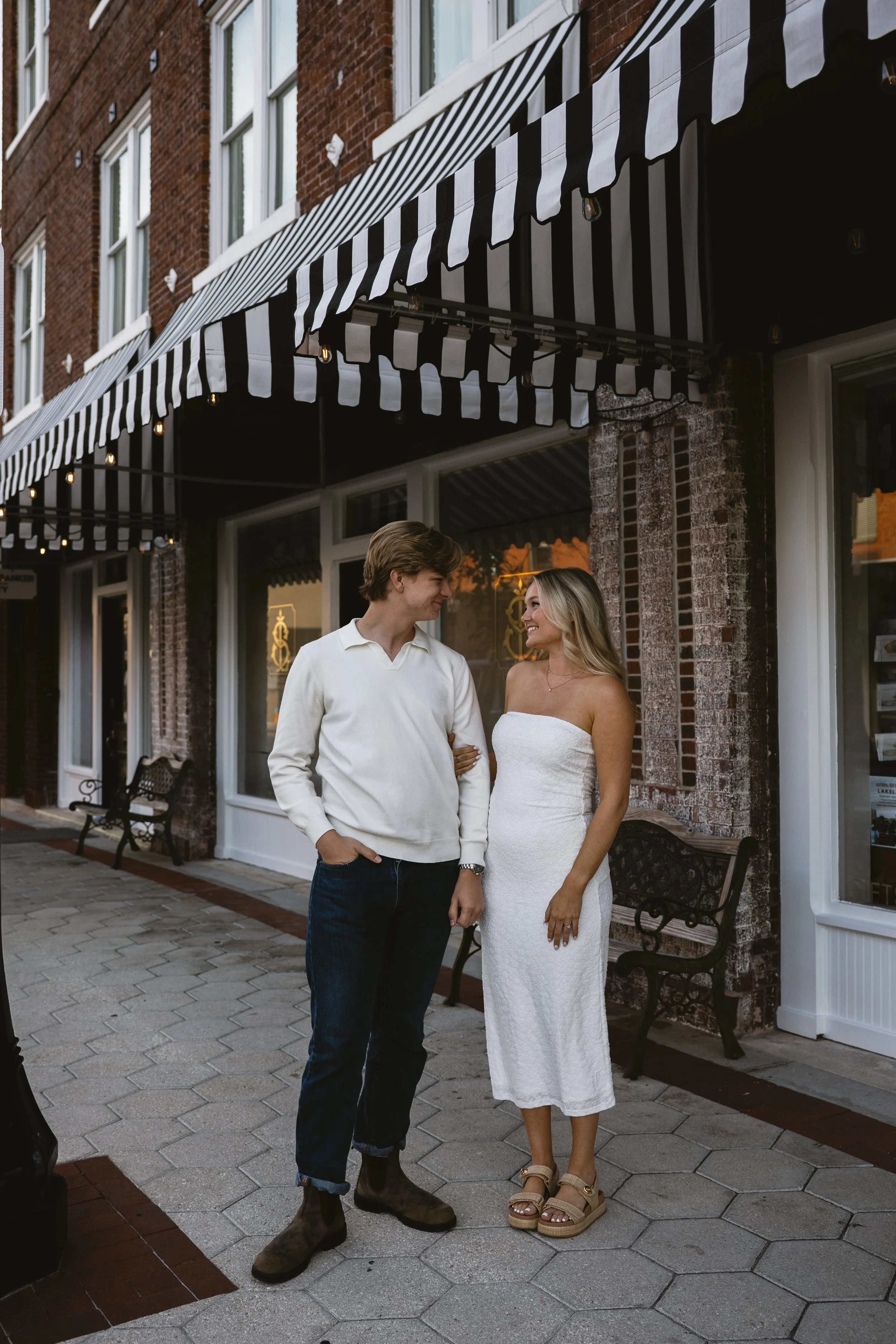 A young couple, a man and a woman, standing outside a brick building with a black and white striped awning, smiling at each other. The man is wearing a white long-sleeve shirt and dark jeans with boots, and the woman is in a strapless white dress wit