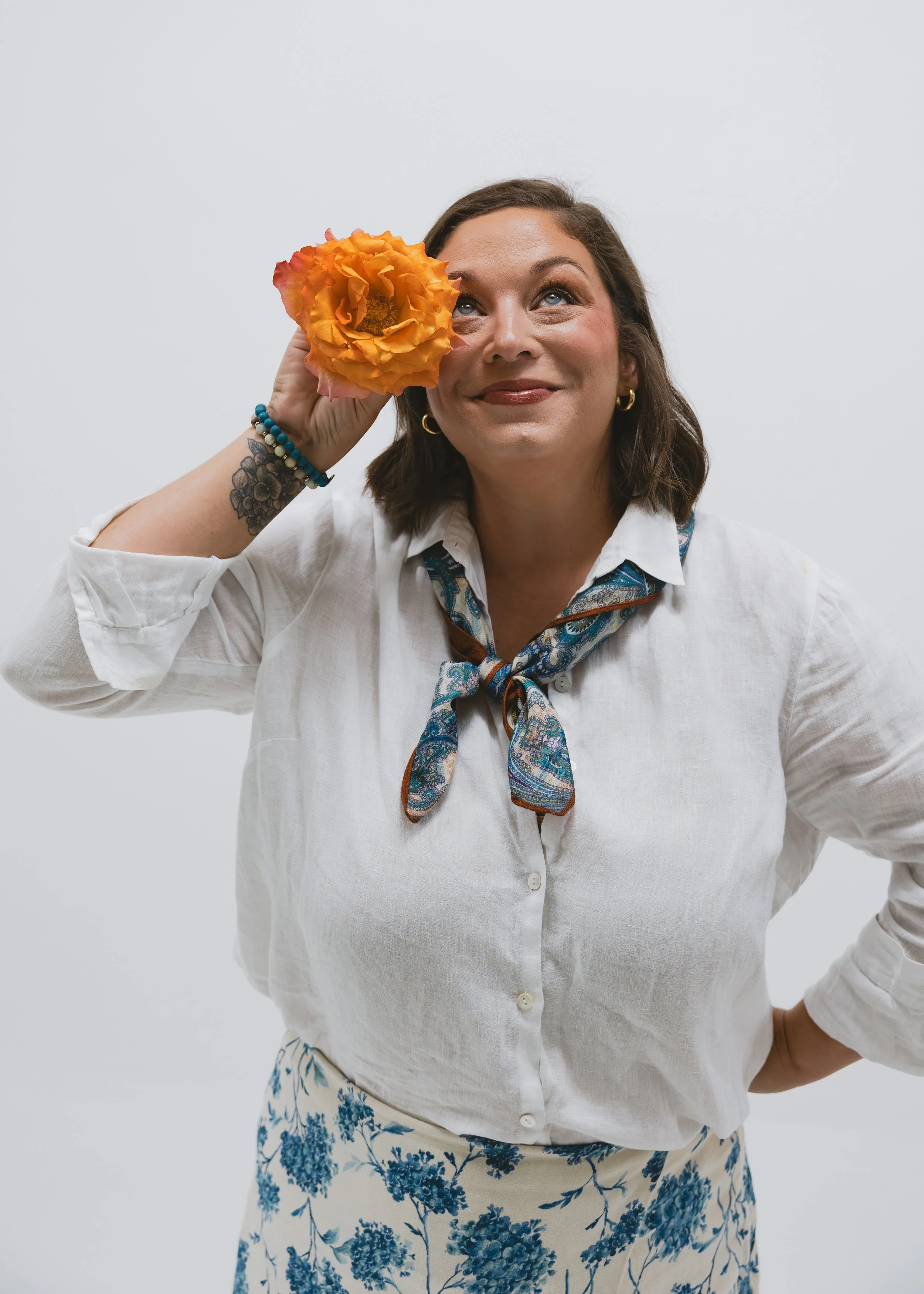 A woman with shoulder-length brown hair, wearing a white blouse with a colorful neck scarf, smiling, holding an orange flower to her face, standing against a plain white background.