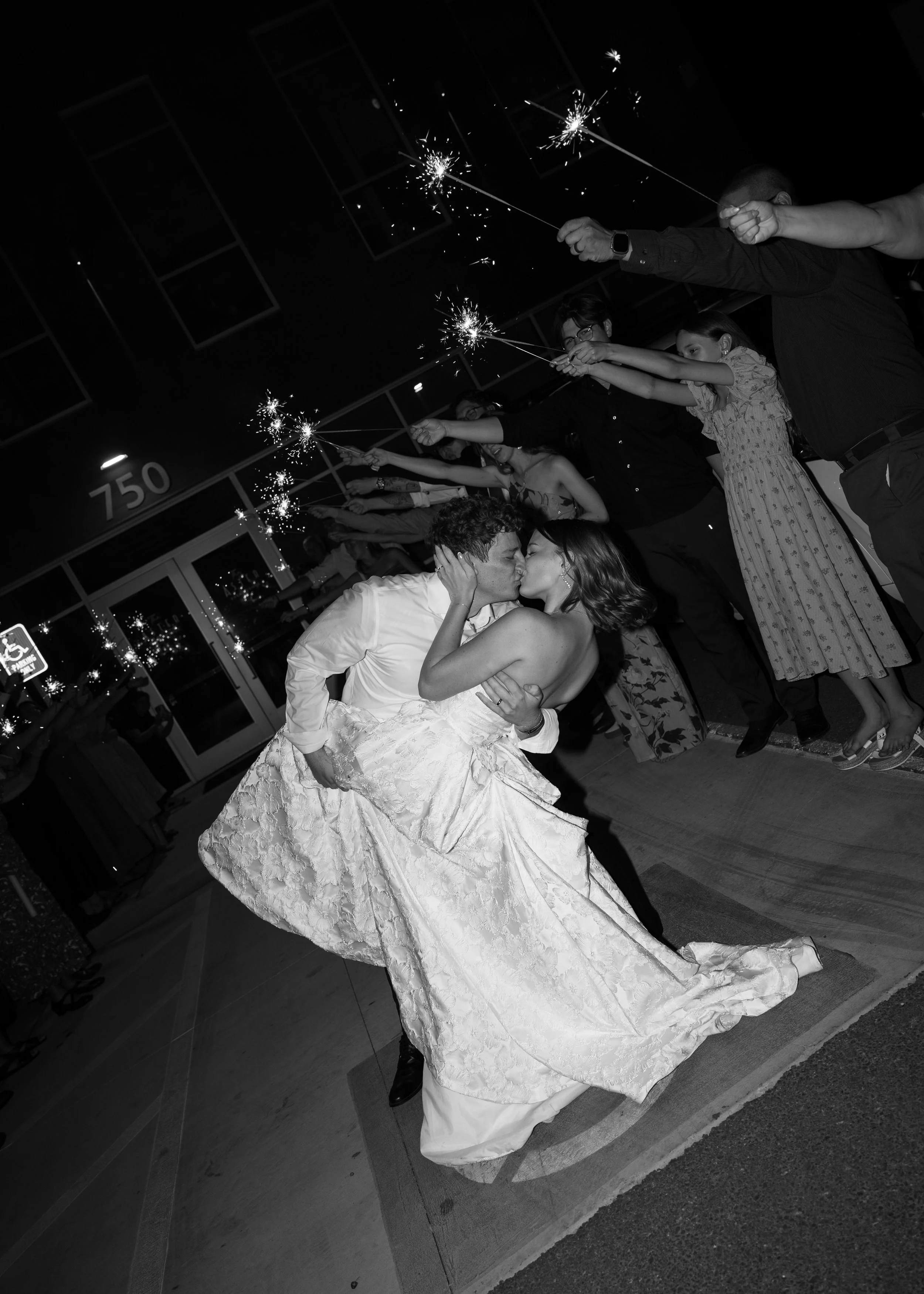 Couple kissing while dancing at a celebration, surrounded by people holding sparklers in a dark setting.