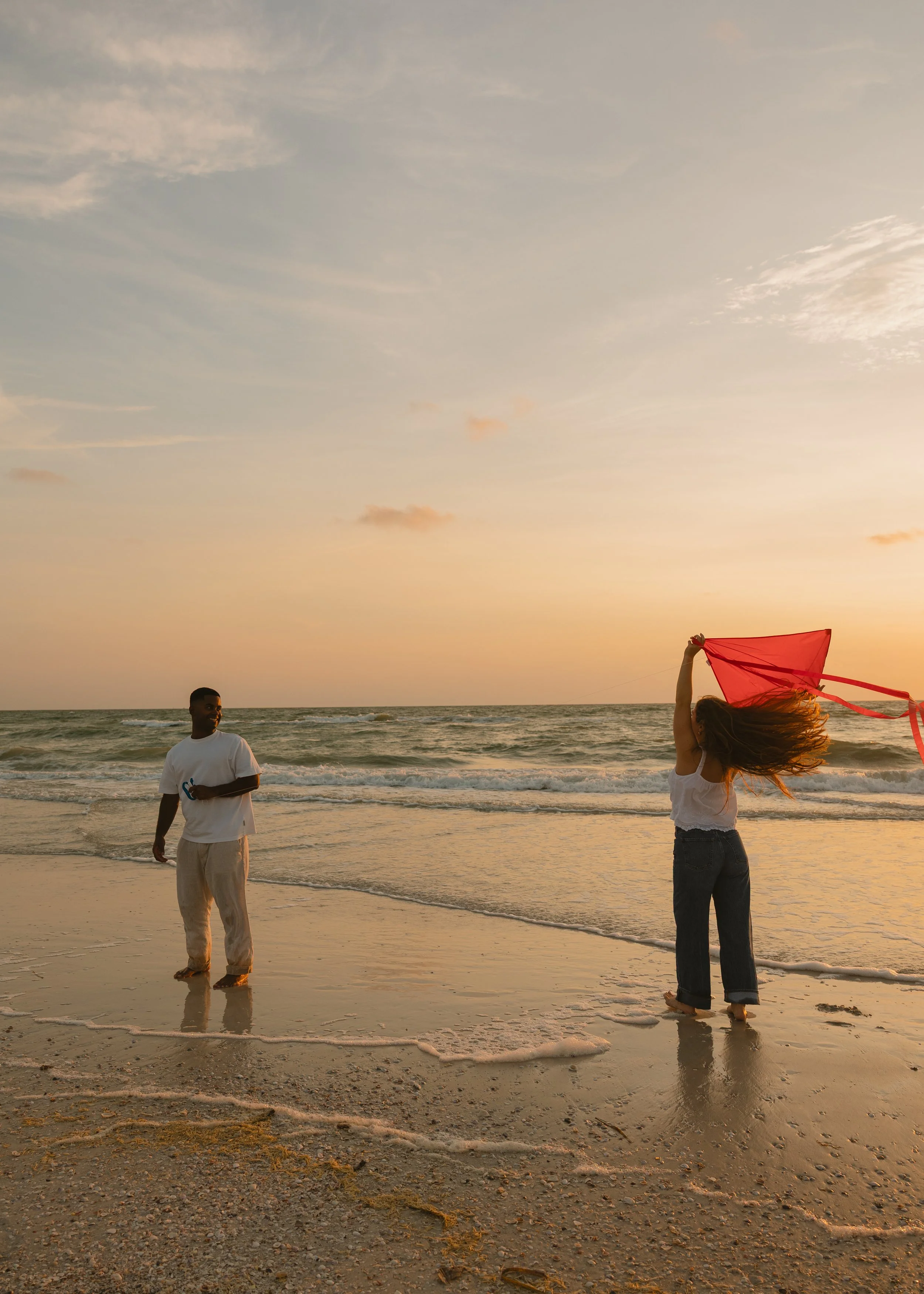 Two people on a beach at sunset. One person is standing in the water, smiling and wearing a white shirt, while the other person, with long hair, is holding a red cloth or scarf and spinning it in the air.