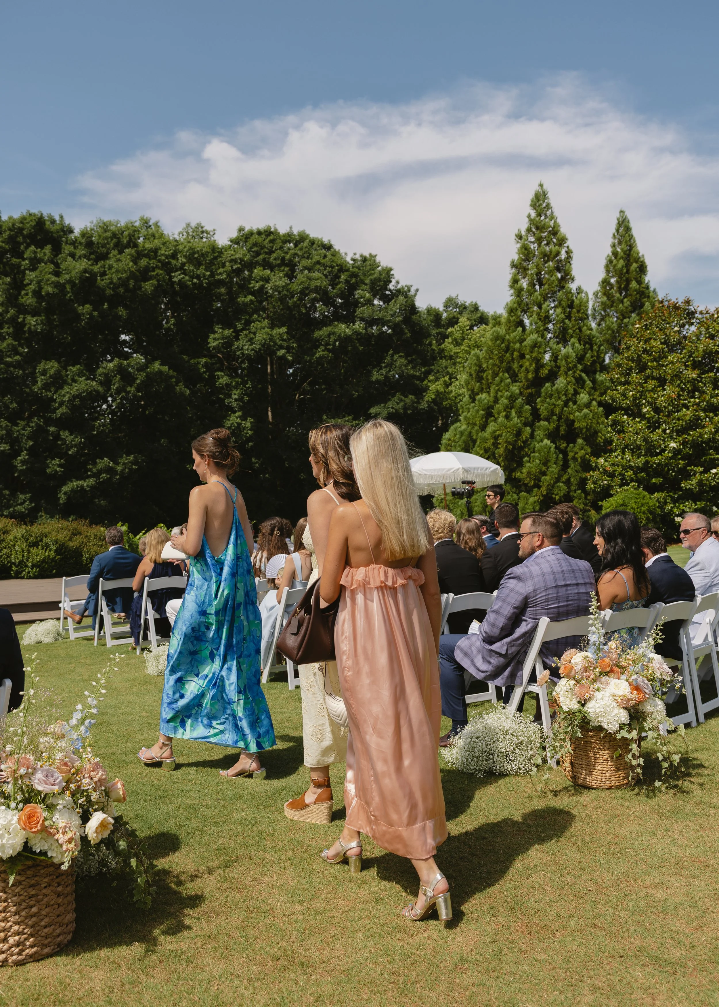 People attending an outdoor wedding ceremony, sitting on white chairs arranged in lawn with flowers and greenery, under a partly cloudy sky.