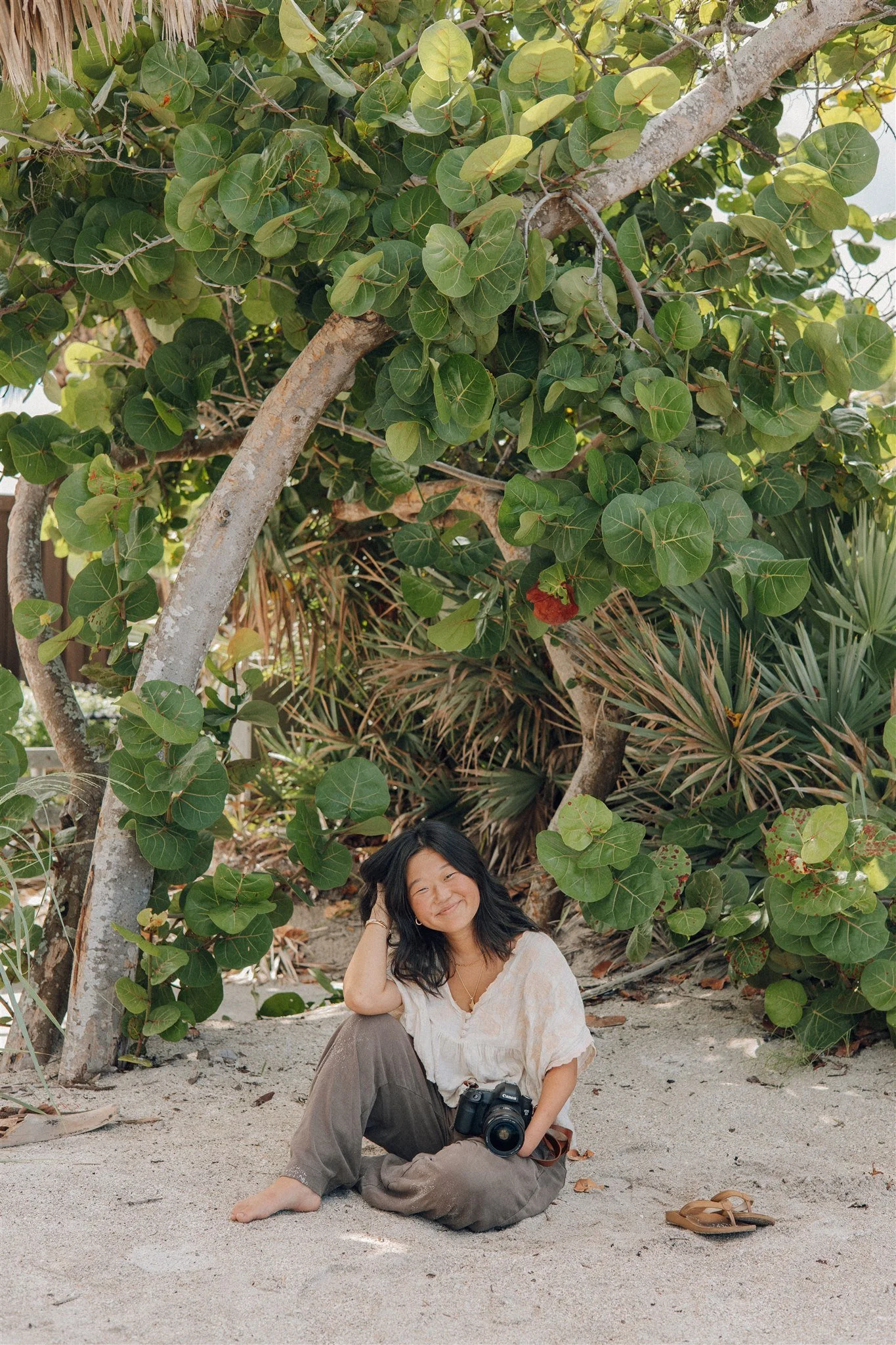 A woman smiling and sitting on sandy ground under a leafy tree, holding a camera, with a pair of sandals nearby.