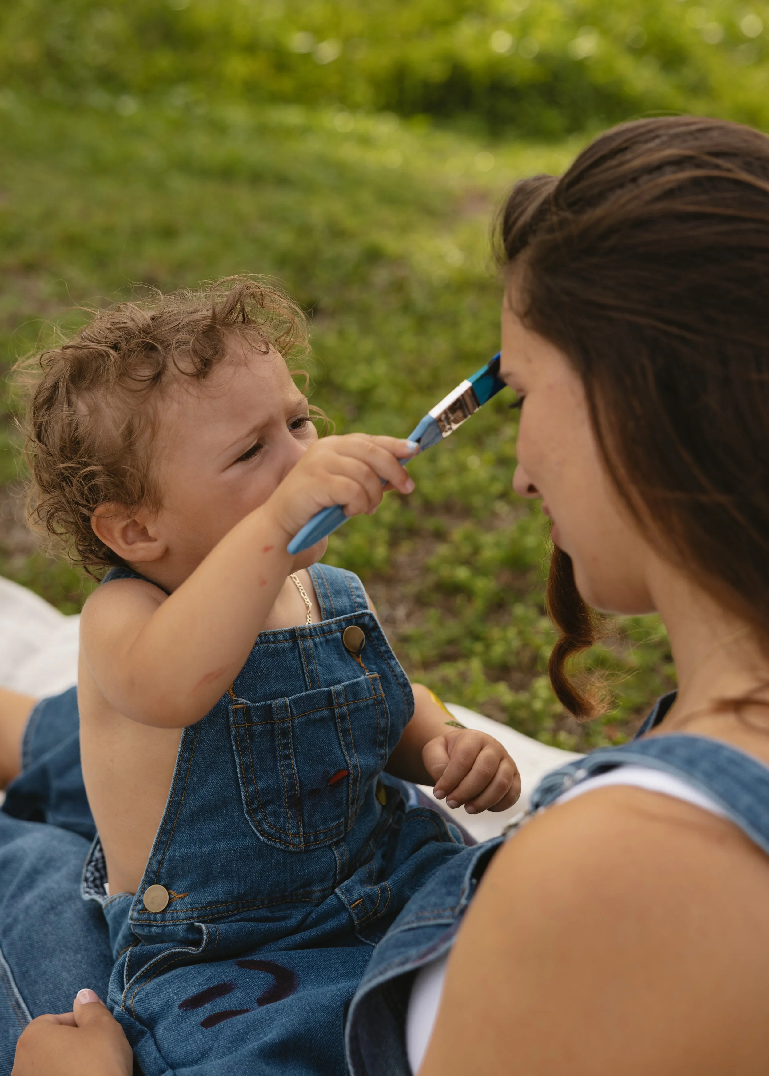 A young child with curly hair and a woman with long dark hair are outdoors on a grassy area. The child is holding a pen and gently touching the woman's forehead, and they are both engaged in a playful or caring moment.