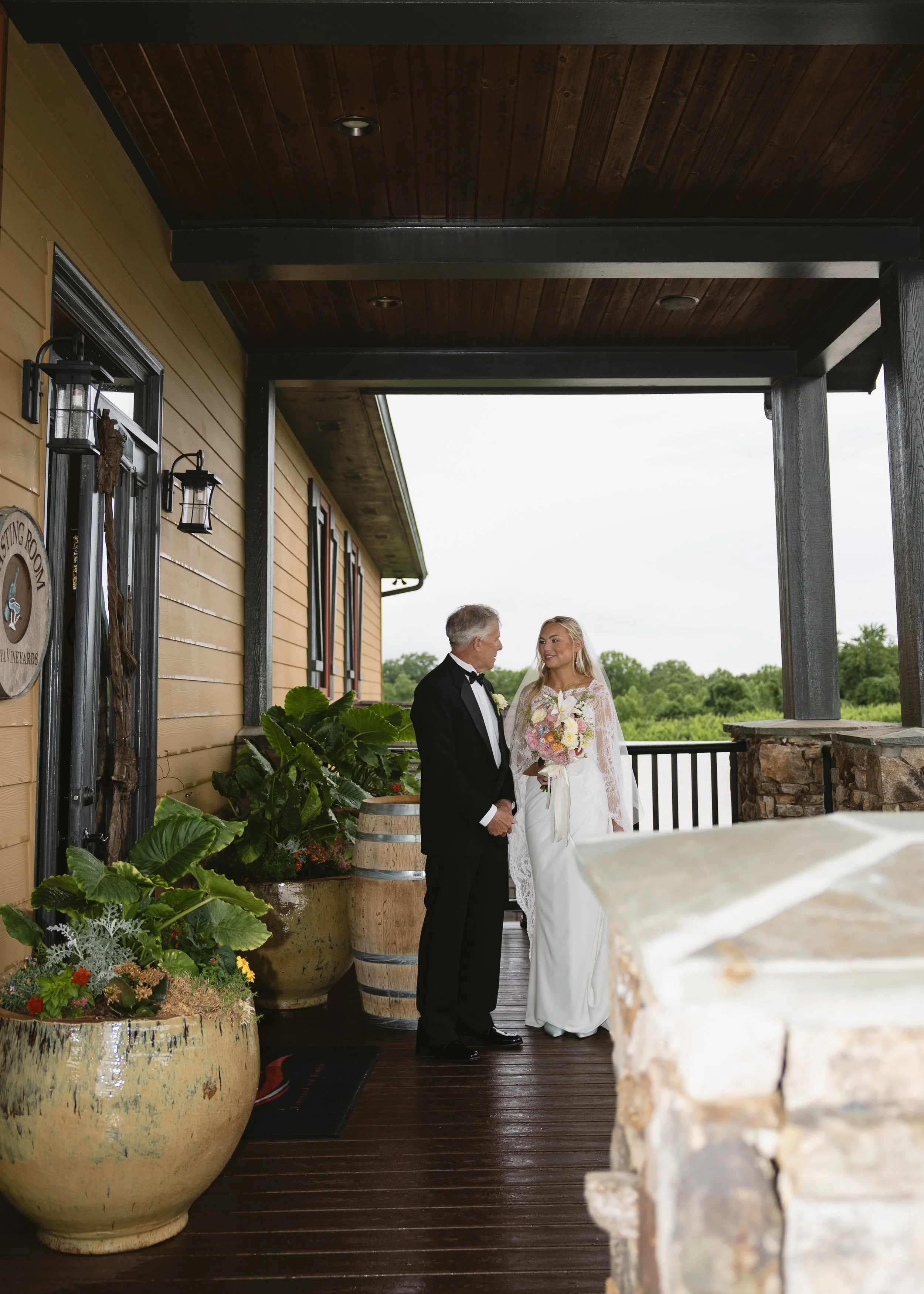 A bride and groom standing on a porch or balcony area. The groom is wearing a black tuxedo, and the bride is in a white wedding dress holding a bouquet of flowers. They are facing each other and smiling.