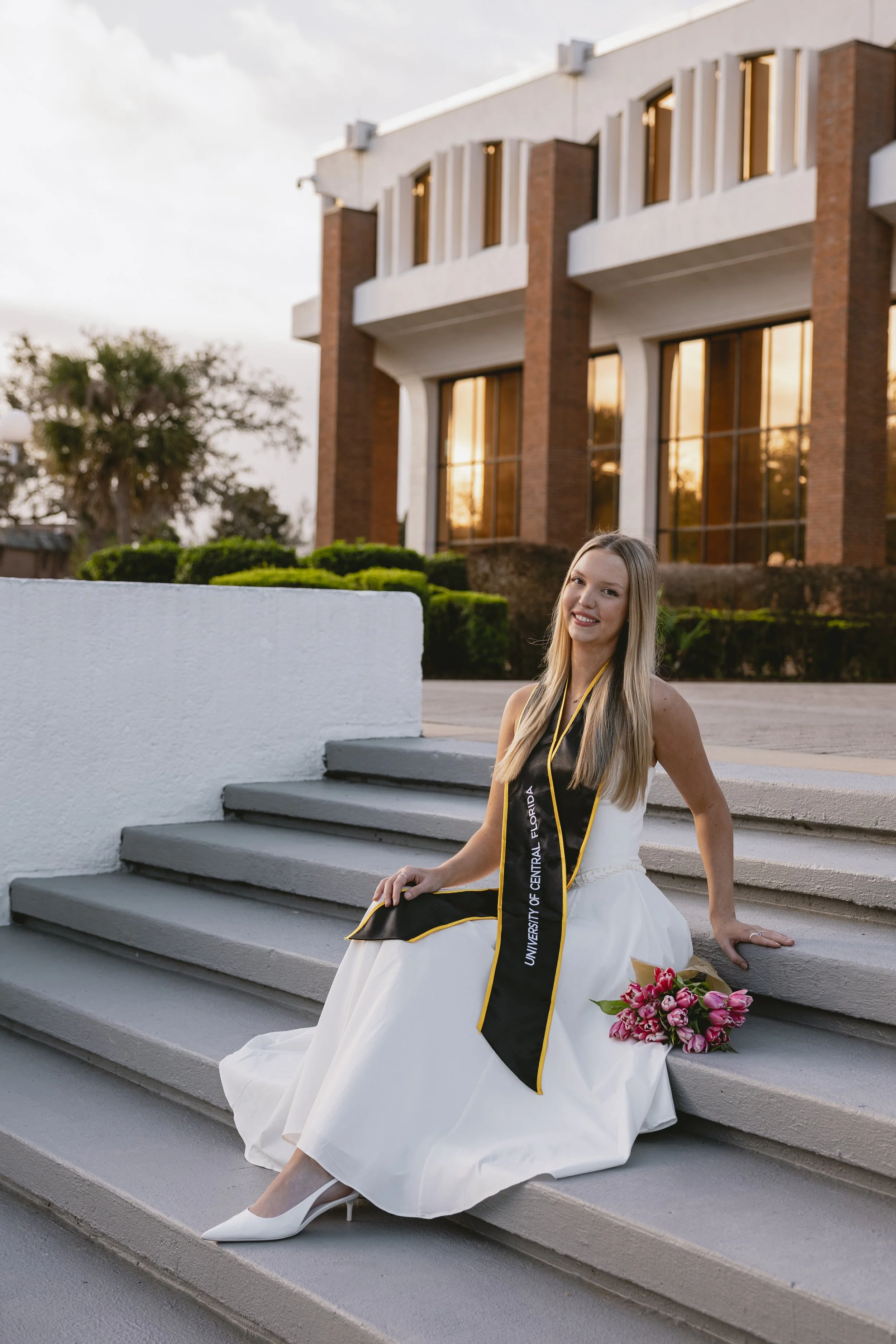 A young woman in a white dress, wearing a black and yellow graduation stole from the University of Central Florida, sitting on steps outside a modern building with large windows during sunset, holding a bouquet of pink flowers, and smiling at the cam