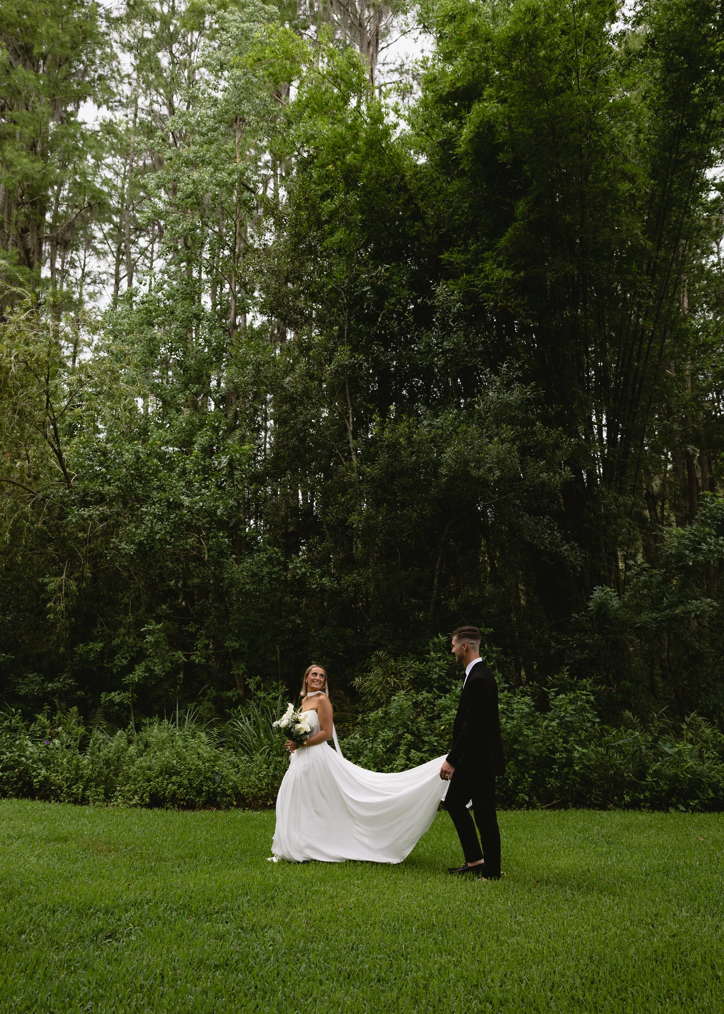 A bride in a white wedding dress holding a bouquet of flowers looking at a groom in a black suit, outdoors on a lush green lawn with tall trees in the background, during daytime.