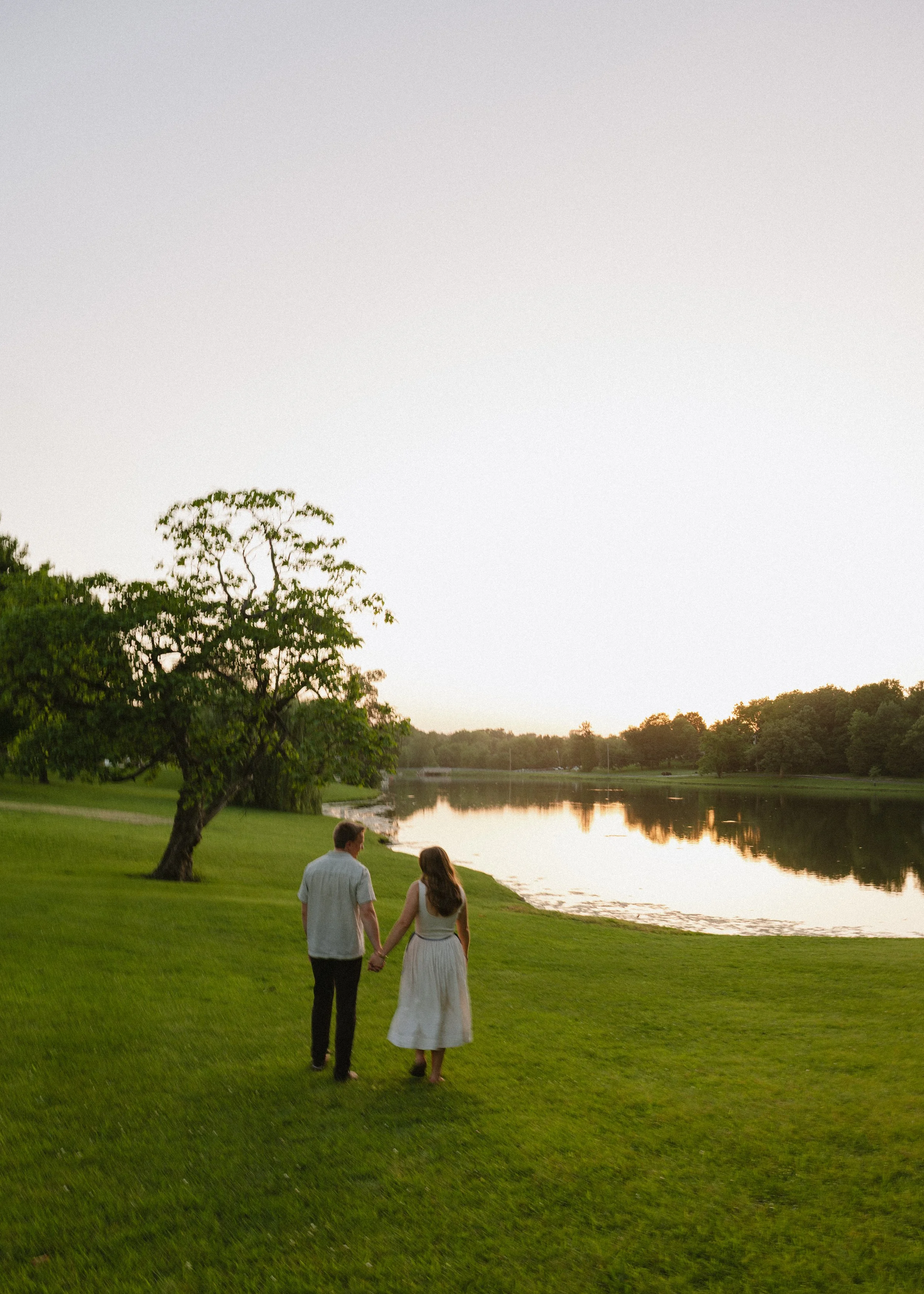 A couple walking hand in hand along the grassy edge of a river during sunset, with trees and calm water reflecting the sky in the background.