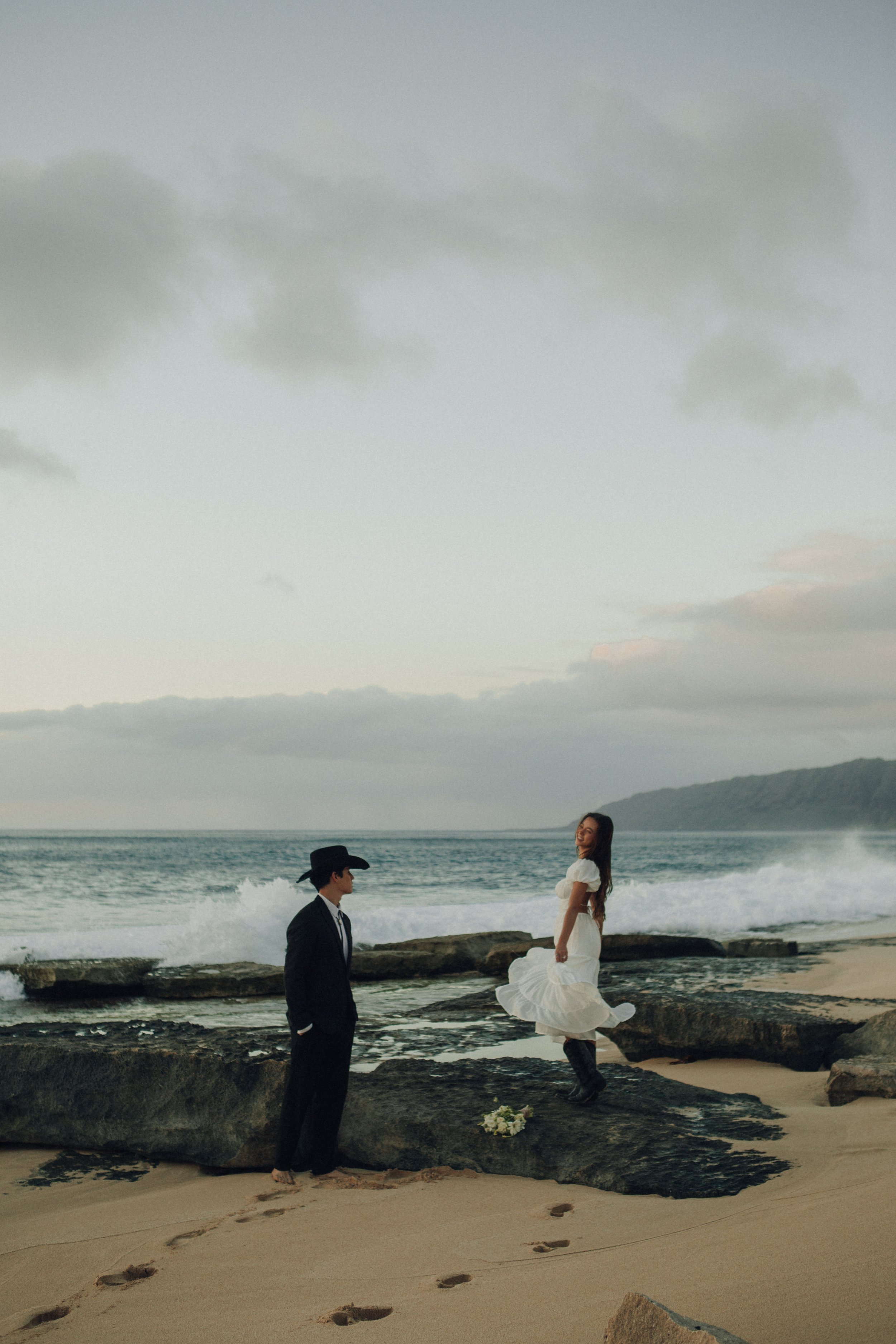 A couple stands on a beach at sunset, with the woman in a white dress and the man in a black suit and cowboy hat. The woman is smiling, and there are flowers on the sand near a rocky area. The ocean waves crash against the rocks, and distant hills ar