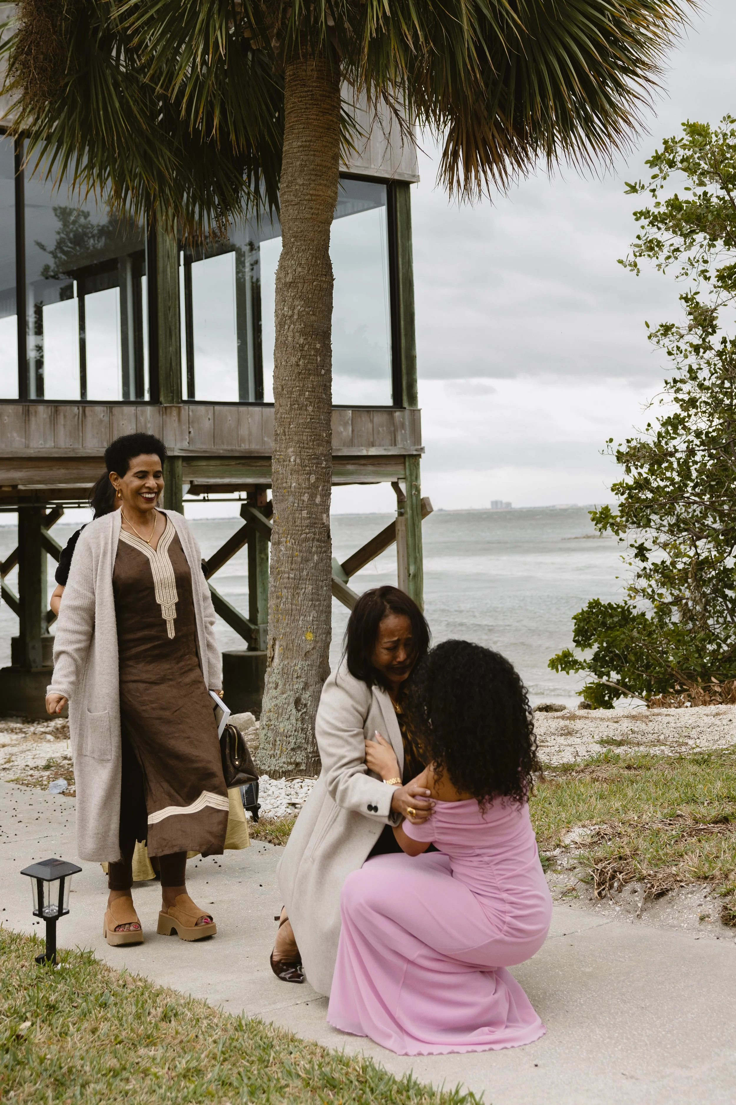 A woman is kneeling and hugging another woman who is seated on the ground near the beach, surrounded by three other women, with a palm tree and a house on stilts in the background.