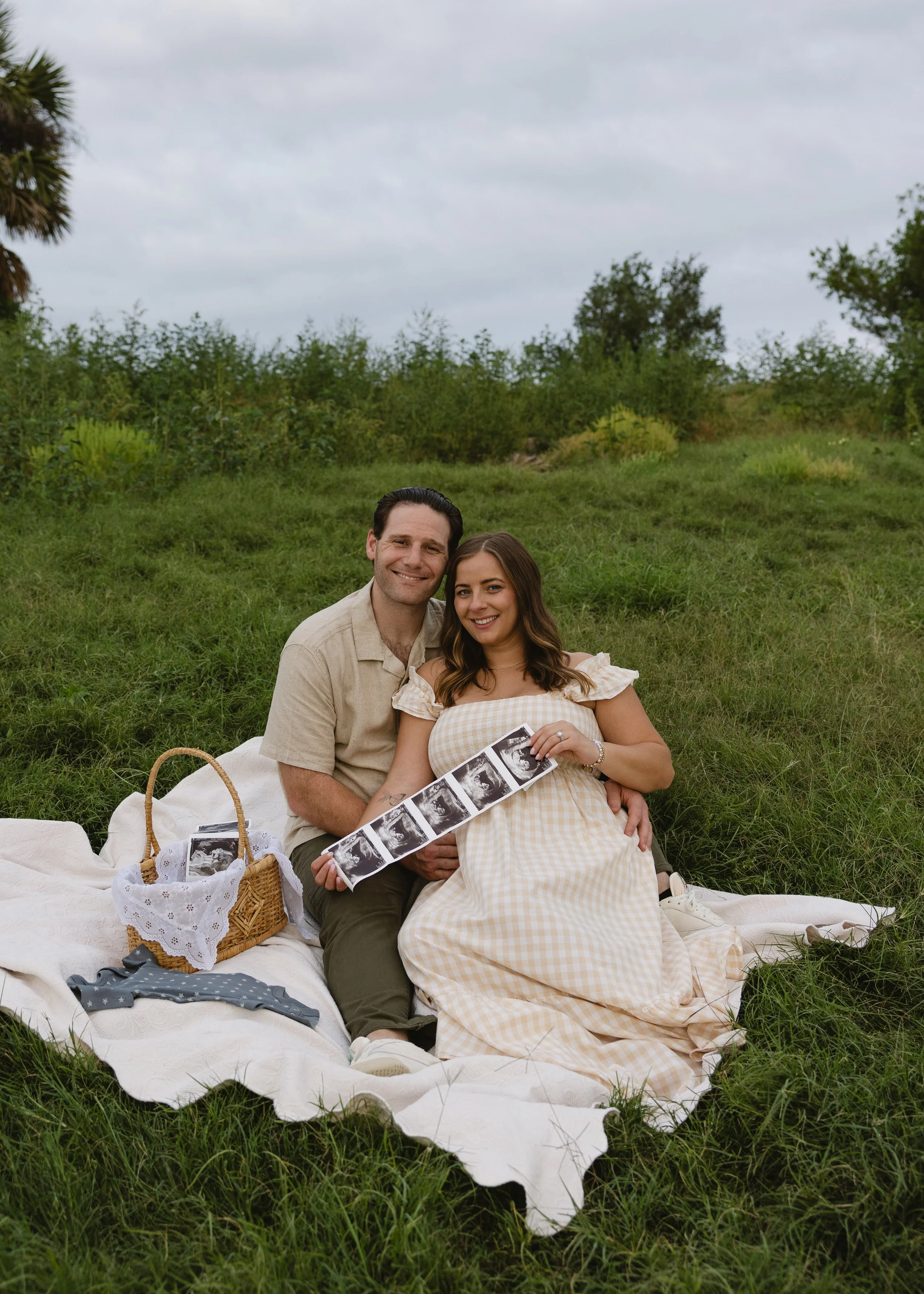 A couple sitting on a picnic blanket in a grassy field, holding a sonogram photo, with a picnic basket and baby clothing nearby, overcast sky in the background.